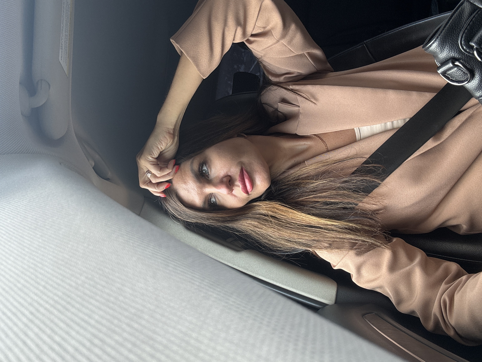 In the city of Chaikovsky, Russia, a woman with long, brown hair and red fingernails is seated in the passenger seat of a car. She is looking out the window with her right hand resting on her head, and a ring is visible on her finger. She is wearing a beige blazer over a light-colored top and a gold necklace. A black seatbelt is fastened across her chest, and a black purse is visible to her right. The interior of the car, including a sun visor and a door handle, is also visible. The lighting suggests it is daytime, with sunlight filtering through the window.