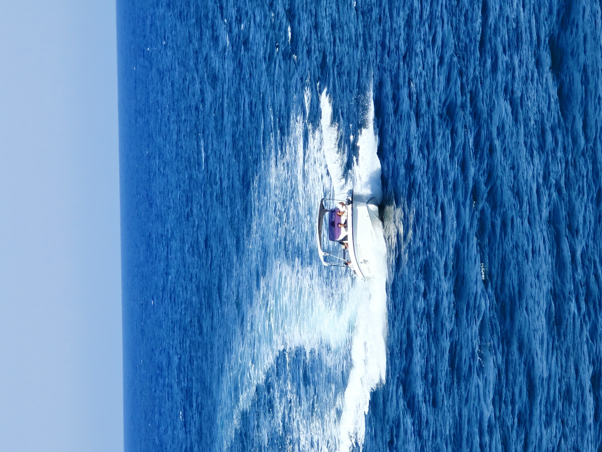 A white motorboat with a canopy is moving through the deep blue sea, creating a white wake behind it. There are three people visible on the boat, two seated and one standing, appearing to be enjoying the ride. The water is choppy with small waves, and the sky is a clear, pale blue. The image captures a sunny day on the water, with the boat cutting through the ocean in a motion that suggests speed and exhilaration. The specific location in Chania, Greece cannot be determined from the image alone.
