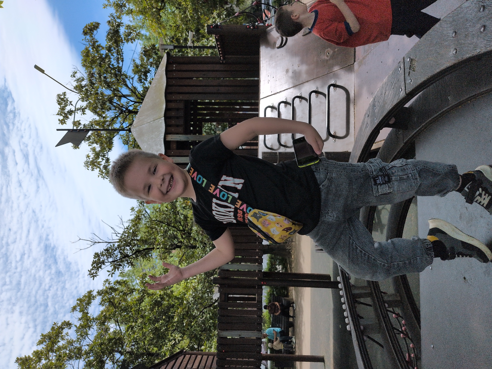 A boy stands on a circular platform, his arm outstretched, smiling and looking directly at the camera. He is dressed in a black T-shirt, gray jeans, and sneakers. He carries a yellow bag around his waist and holds a mobile phone in his hand. Another boy is seen lying on a surface behind him, wearing a red shirt. In the background, there is a wooden structure resembling a playground and people seated. Trees and the sky can be seen. The location appears to be Sochi, Russia.