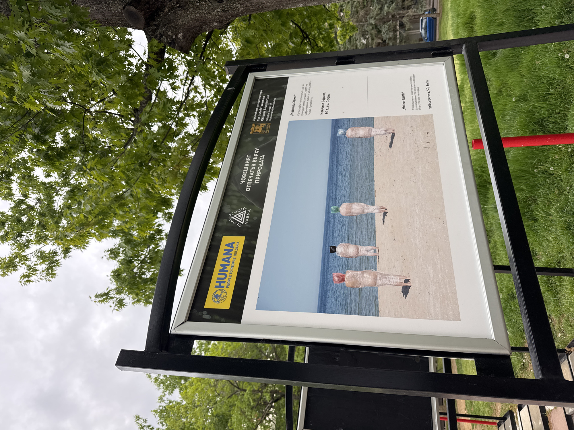 The image shows a sign in what appears to be a park in Blagoevgrad, Bulgaria. The sign is black with a white framed poster. The poster features an image of four figures standing on a beach, each wrapped in transparent material with a different colored top (blue, green, black, and red). The backdrop is the sea and sky. Text on the poster in Bulgarian and English identifies the artwork as "Mother Earth" by Ivelina Berova from Sofia, age 50. The text explains that the photographer raises the question of the boundary between modern life's conveniences and the price nature pays. Logos on the sign include Humana People to People Bulgaria and Dedal School Varna. Text on the sign indicates the exhibition is organized with the support of the Municipality of Blagoevgrad. The sign is surrounded by grass and trees. A portion of a red pole and other foliage are visible in the background.