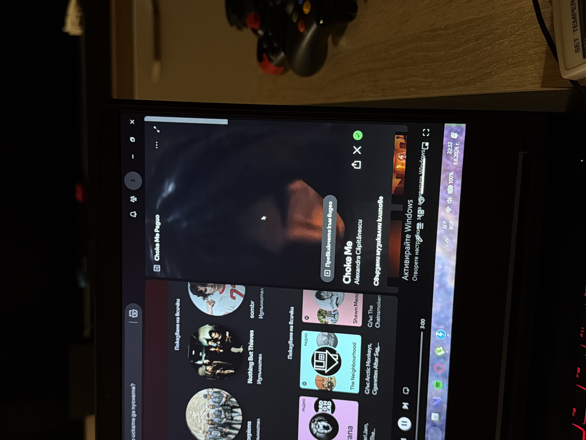 The image shows a laptop screen displaying a Spotify interface. The Spotify application features album art and song titles in what appears to be Bulgarian language. The active song displayed is "Choke Me" by Alexandra Căpitănescu. In the top right corner of the screen, there is a notification indicating "3" people are in a group, and further to the right, window control buttons (minimize, maximize, close). The bottom taskbar of the operating system shows icons for Windows, Microsoft Store, Spotify, and other applications, along with the date "9.6.2026" and time "22:32". The laptop is placed on a wooden surface, possibly a desk. To the right of the laptop, a video game controller is visible, with its buttons and joysticks clearly discernible. The overall lighting suggests an indoor environment, possibly at night, with the screen being the primary light source. There is no discernible information in the image to suggest a specific location within Sofia, Bulgaria.