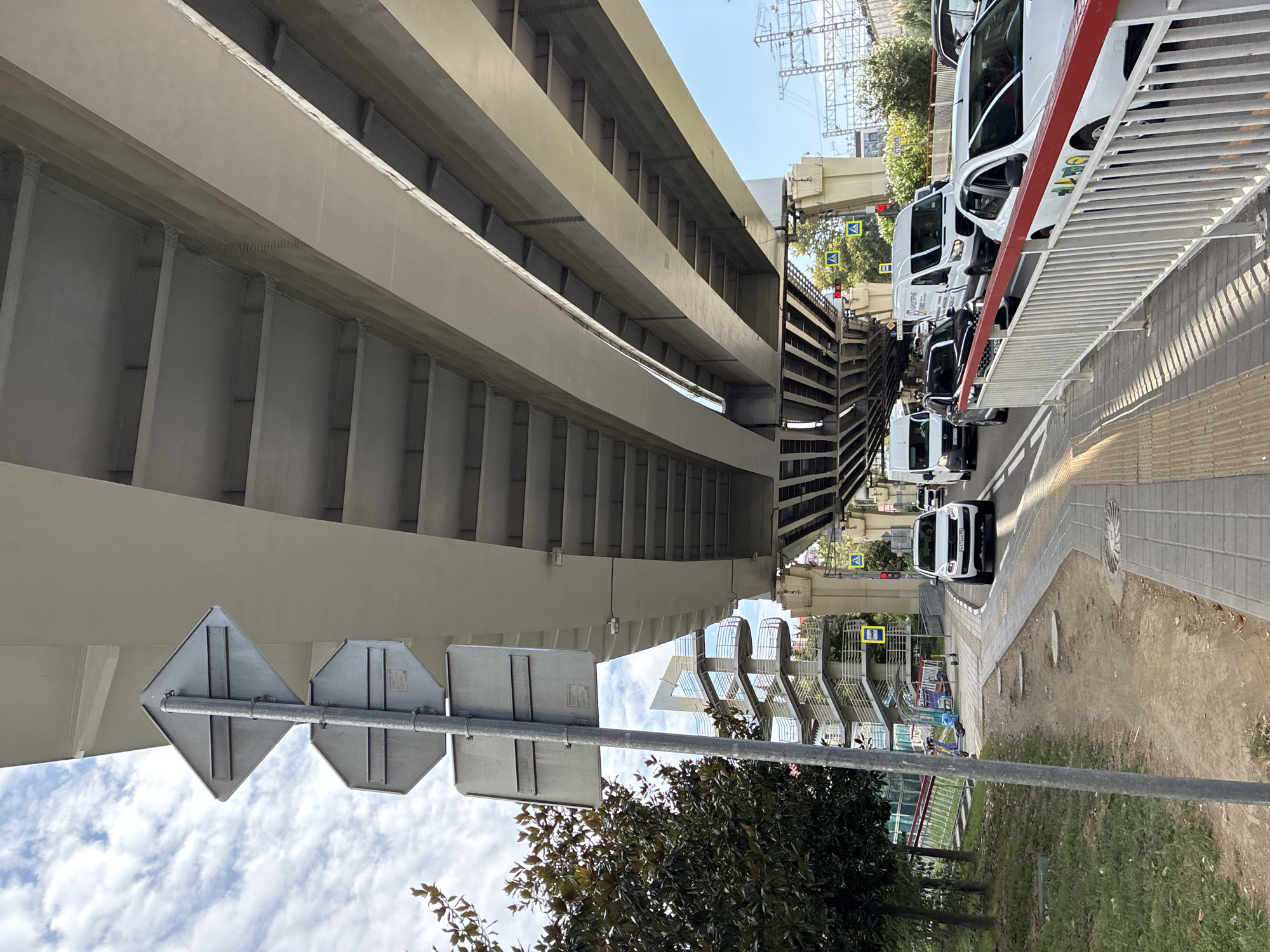 The image shows a street scene in Sochi, Russia, under an elevated overpass. A white SUV is in the foreground, driving on the right side of the road. Several other vehicles, including vans and a black car, are also visible on the road, moving in the same direction. Pedestrian sidewalks and a grassy area with trees are present on the left side of the image. A modern multi-level pedestrian bridge with ramps and staircases is visible behind the trees. Traffic signs are mounted on a pole near the sidewalk. The sky is partly cloudy.