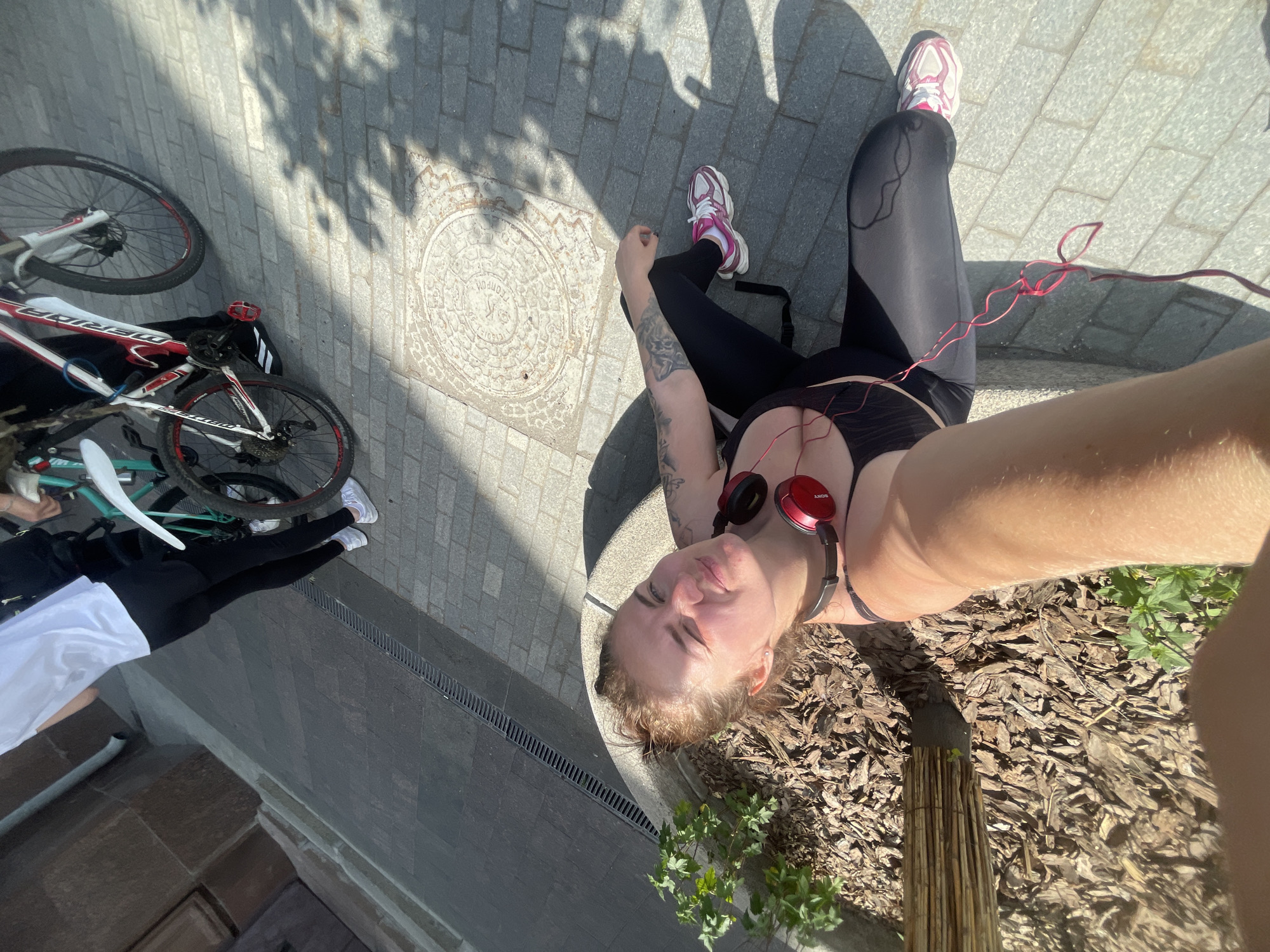 A high-angle, medium shot depicts a woman lying on a paved sidewalk next to a curb in Moscow, Russia. The woman is wearing black leggings and a black top. She has tattoos on her left arm and is wearing red headphones. A red cable extends from the headphones towards her leg. She is lying next to an area covered with wood mulch and small plants. A bicycle with a red and white frame lies on the sidewalk next to another person wearing white shoes and black pants. The scene is well-lit, suggesting it is daytime.