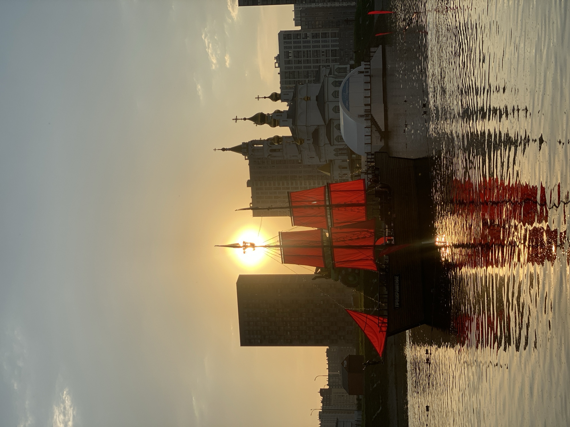 The image shows a sunset scene over a body of water, likely a river or lake, in Yekaterinburg, Russia. In the foreground, there is a ship with red sails. The reflection of the ship is visible in the water. In the background, there is the Church on Blood in Honour of All Saints Resplendent in the Russian Land and modern apartment buildings. The sun is directly behind the ship.