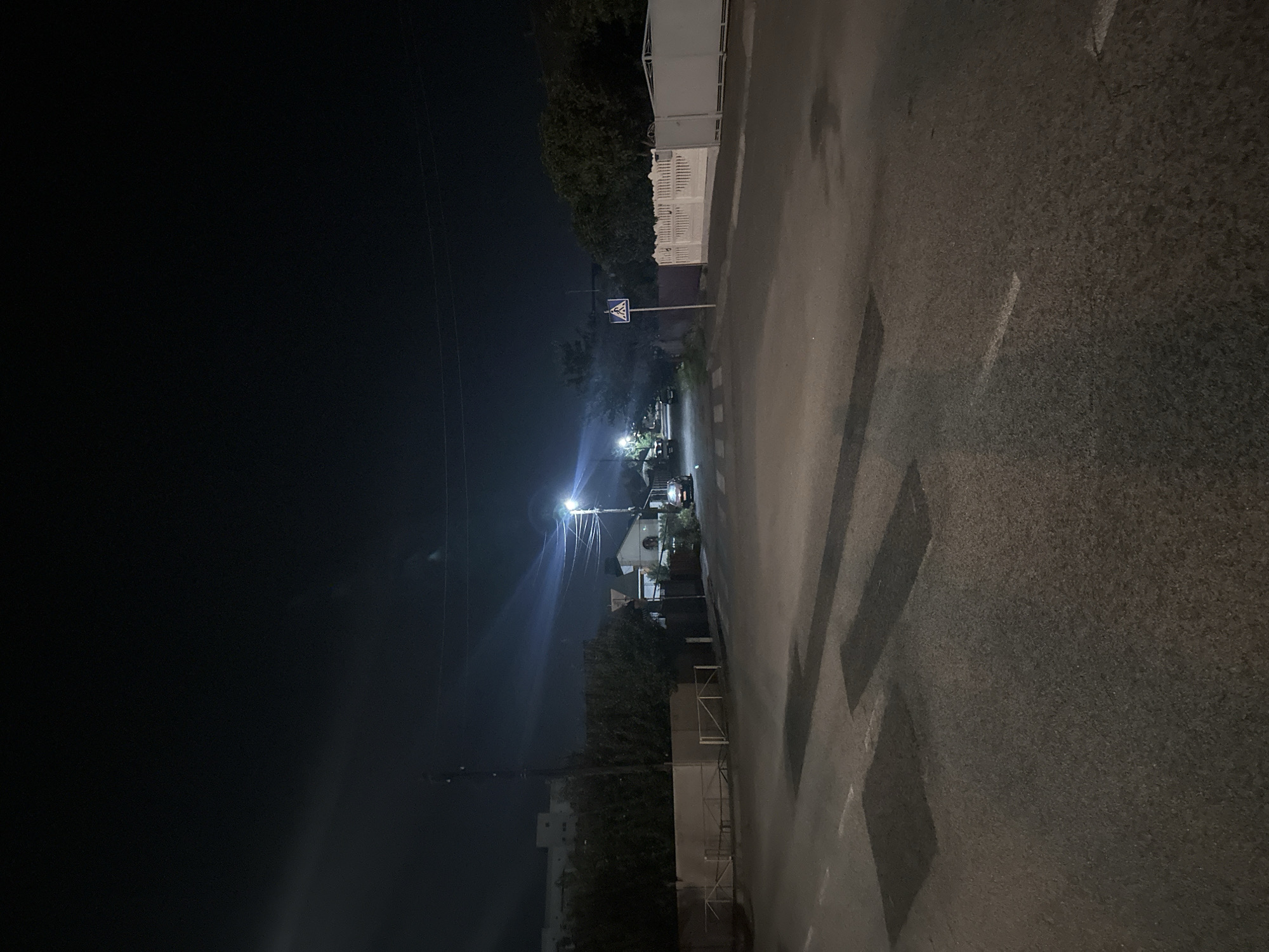 The image depicts a street scene at night in Khmilnyk, Ukraine. The foreground shows an asphalt road with painted crosswalk markings and dark, rectangular patches, possibly indicating repairs or faded markings. The road curves slightly to the right. On the left side of the road, there is a low, light-colored fence topped with metal railings. Behind the fence, trees and the tops of buildings are visible. A utility pole with wires stretches across the scene. To the right, a pedestrian crossing sign is visible. Beyond the crossing, there is a white, ornate fence and a gate. Further back, houses with illuminated windows and a street light casting a bright glare are present. A car is parked on the right side of the road. The sky is dark with some light streaks, possibly from atmospheric conditions or light pollution.