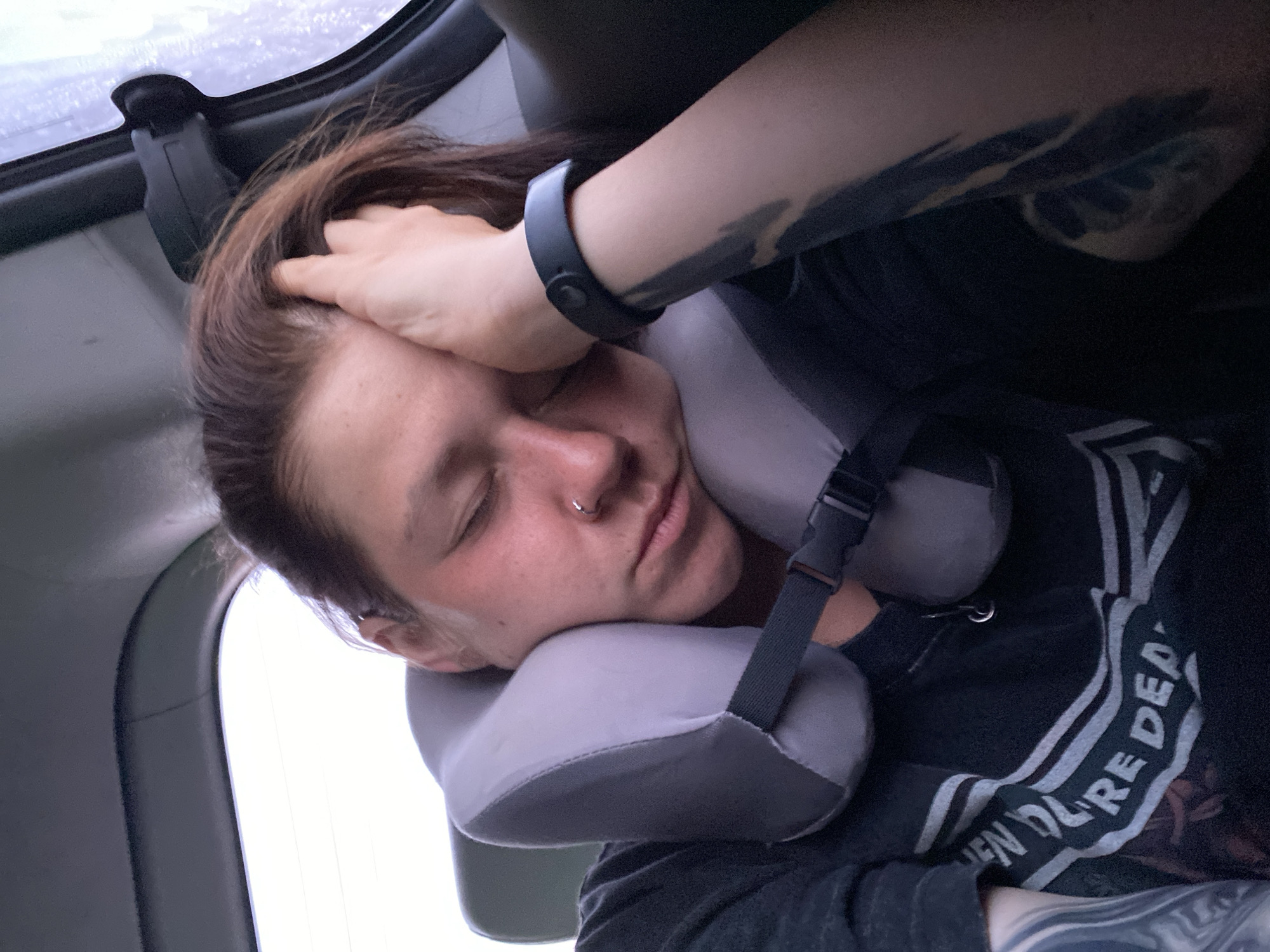 The image shows a person sleeping in the back of a vehicle, likely a car, indicated by the headrest and window. The person is wearing a dark t-shirt with white lettering that is partially visible, reading "EN YOU'RE DEA". They are resting their head on a gray neck pillow, which is secured with a black strap. The person has a nose ring and visible tattoos on their arm. The lighting suggests it is daytime, but the external environment is not clearly visible. The scene appears to be indoors, within the confines of a moving vehicle. There is no information in the image to suggest a specific location in the city of Novyi Kal'chir, Russia.