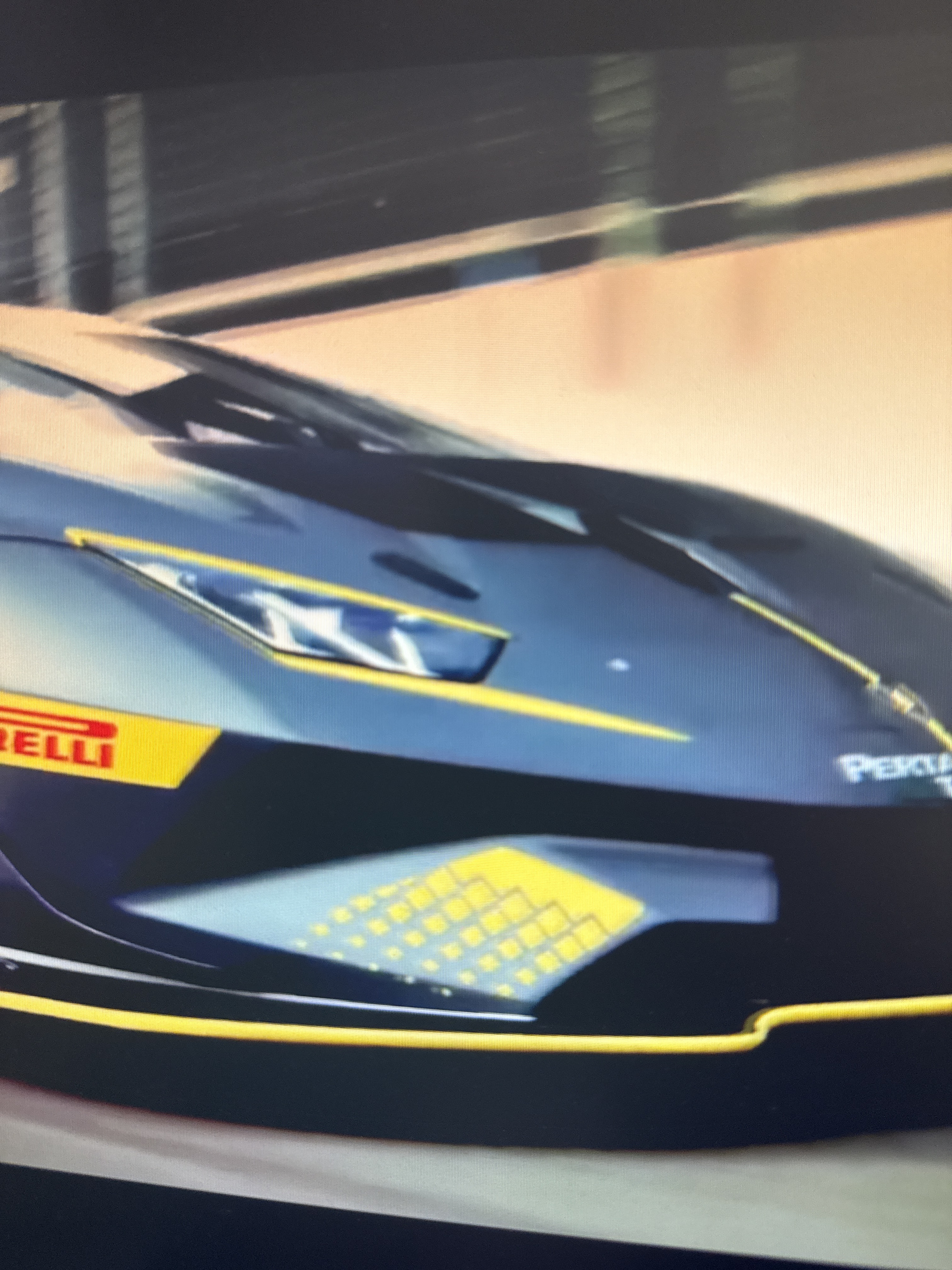 The image shows a close-up, slightly blurry view of the front of a dark blue and yellow race car. The car has yellow accents and a yellow stripe with the word "IRELLI" in red visible on the side. There is also a logo that appears to say "PERTA" on the front right of the car. The background is out of focus, but suggests an indoor setting, possibly a garage or pit area, with horizontal lines that could be part of a building structure. The lighting appears to be artificial. The image does not provide any information about the city of Netanya, Israel, or any specific location. There are no people visible in the image.