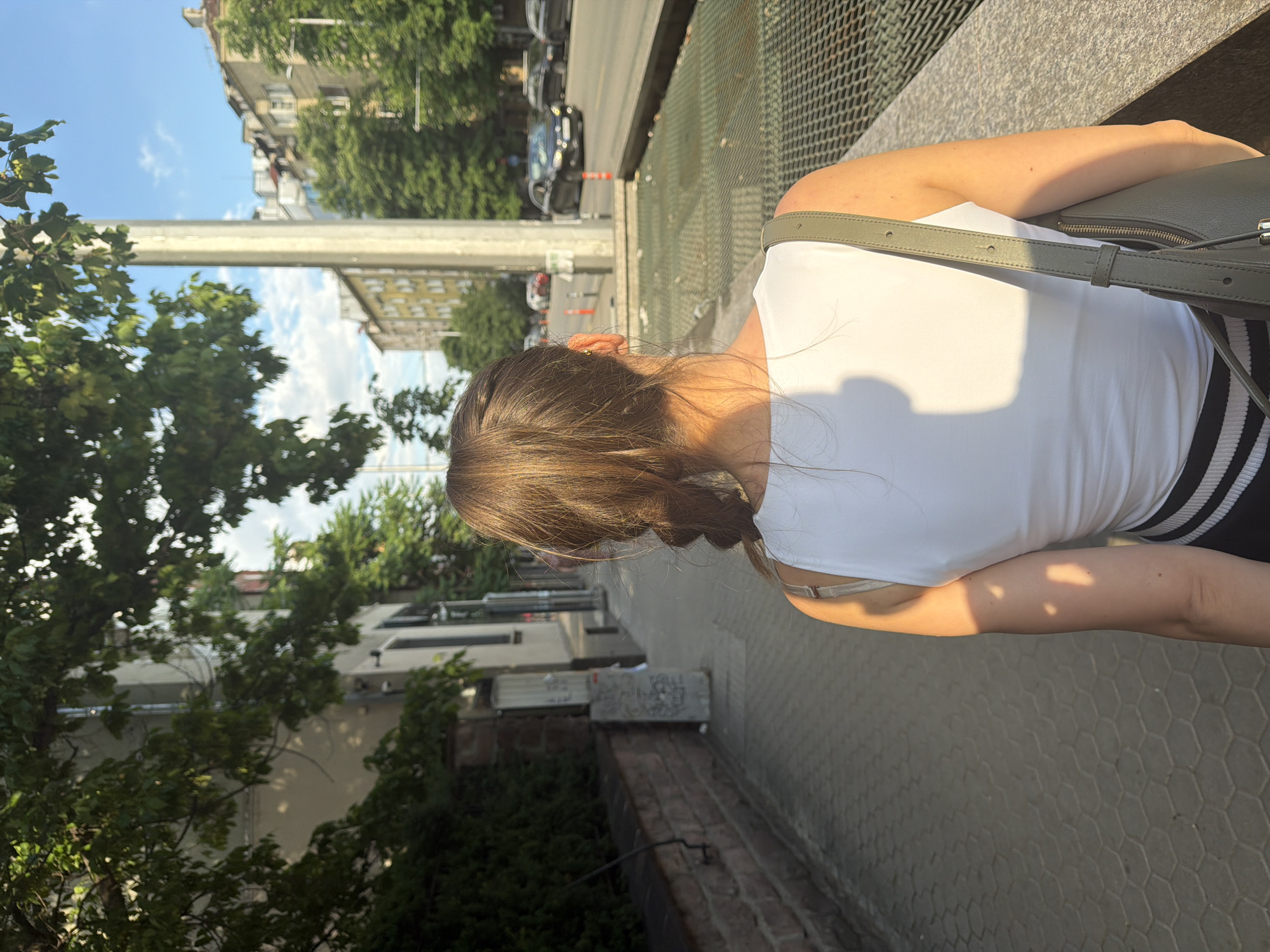 The image depicts an urban street scene in Sofia, Bulgaria, captured from a rear perspective, focusing on a person walking away from the camera on a sunlit sidewalk.
A person, appearing female, with brown hair styled in a single braid, is centered in the midground. She wears a white sleeveless top and black and white horizontally striped bottoms. A grey/olive-green shoulder bag is slung over her right shoulder. Sunlight casts prominent shadows from her body and nearby foliage onto her back and the sidewalk. A small, golden earring is visible on her left earlobe.
The sidewalk, paved with grey hexagonal blocks, extends from the foreground into the distance. To the person's left, a low stone wall is visible, topped with dark green foliage. Behind this, an apartment building with a light-colored facade and visible drainpipes stands partially obscured by trees. A utility box with graffiti is present against the wall.
To the person's right, a concrete barrier with a horizontal metal grate on top parallels the sidewalk. Beyond this barrier, a tall, light-colored utility pole stands vertically. The street is visible further to the right, with asphalt pavement and a black sedan driving away in the distance. Orange traffic bollards are spaced along the curb.
In the background, multi-story residential buildings line both sides of the street under a bright blue sky with scattered white clouds. Lush green trees are abundant, casting dappled shadows across the scene. The overall impression is a sunny day in a busy city environment.