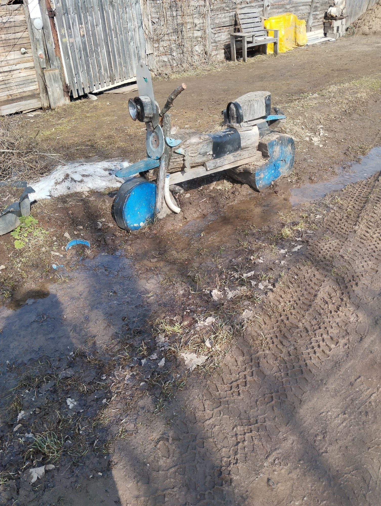 The scene depicts a handmade wooden scooter, painted in blue and black, positioned in a muddy, rural environment. The scooter is constructed from various pieces of wood, appearing weathered and worn. The immediate foreground is dominated by a muddy puddle with tire tracks visible on the right side. The background features a weathered wooden building, a wooden bench, and some yellow-covered objects, possibly hay bales. The location is speculated to be Grishanki, Russia. No people are visible in the image.