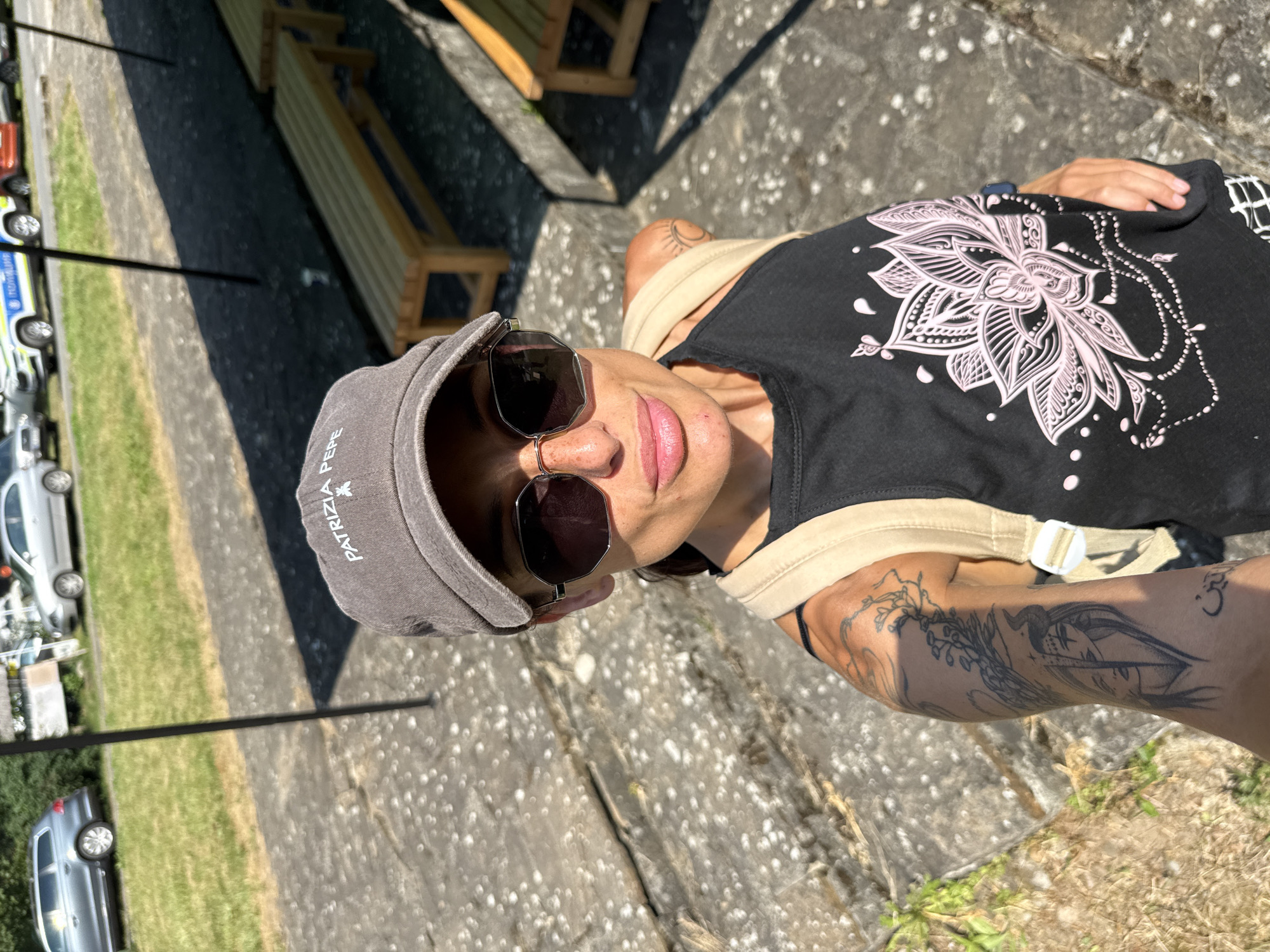 A woman is taking a selfie outdoors. She is wearing a grey baseball cap, sunglasses, and a black tank top with a pink lotus flower design. She has tattoos on her left arm. She is standing on stone steps with wooden benches in the background. Cars and a grassy area are visible behind her. The location is the town of Ribaritsa, Bulgaria.