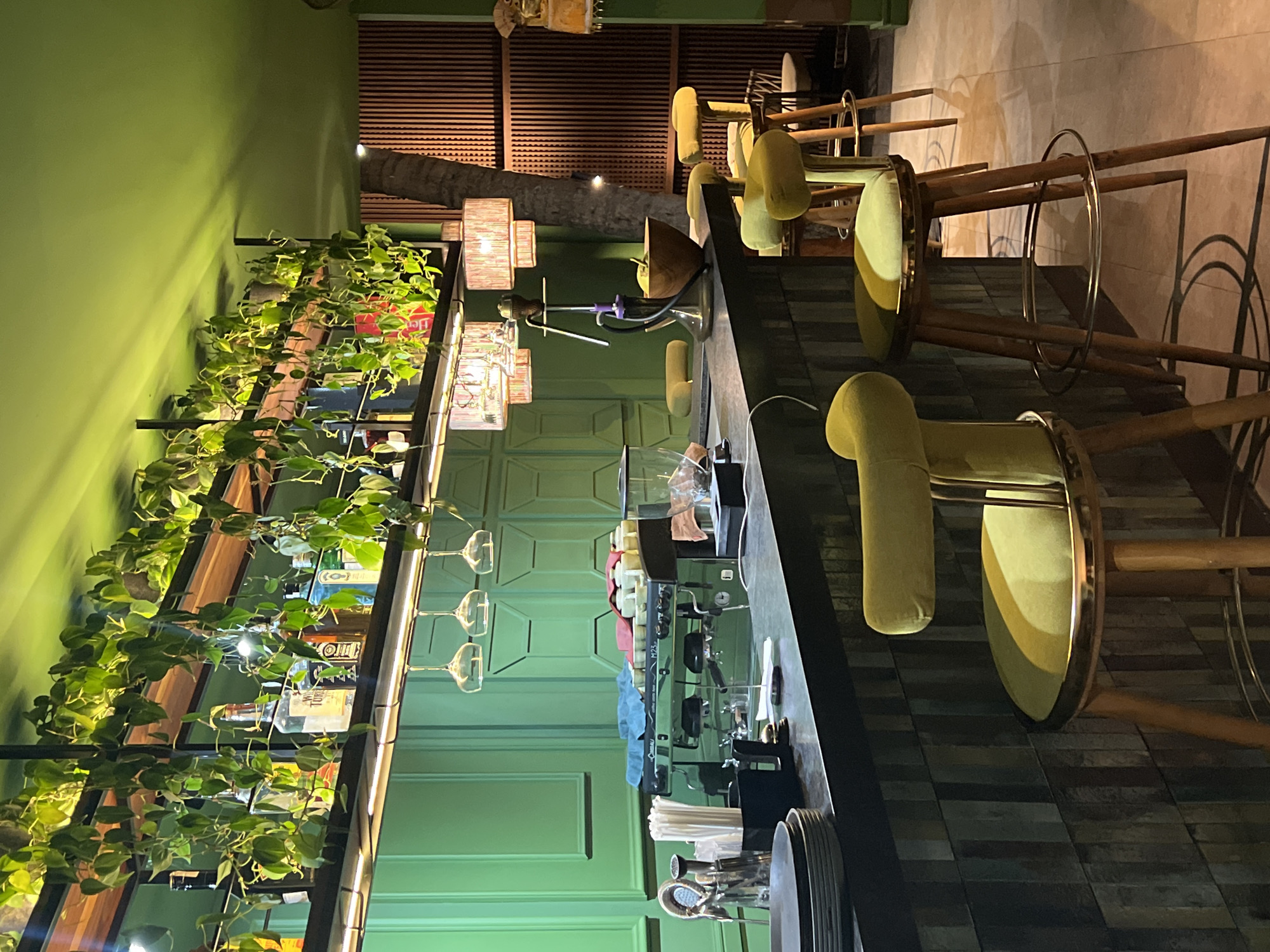 The image shows the interior of a bar or restaurant in Ubud District, Indonesia. The space features a long, dark-colored bar counter with several high-backed yellow bar stools arranged along its length. Behind the bar, there's a green wall with a shelf unit adorned with plants and bottles, suggesting a focus on ambiance and greenery. Above the bar, there are decorative lamps with fabric shades that are mounted to what looks to be a tree trunk. The flooring has a dark, patterned tile. The overall atmosphere appears upscale and stylish.