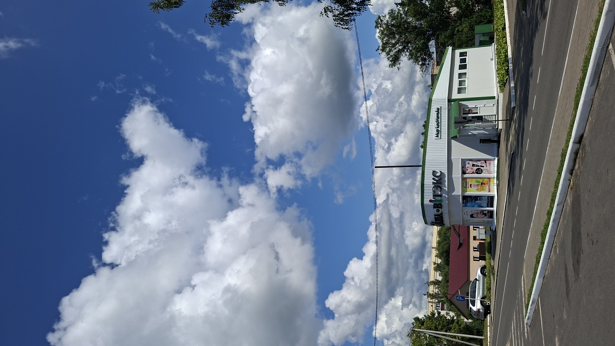 In the city of Myadzyel, Belarus, on a bright, cloud-filled day, a white and green commercial building stands prominently on the right side of the frame. The building features a green awning over its entrance and displays the text "ВИТЭКС" in large white letters on its facade, along with "Мир косметики" to the right. The storefront windows showcase various cosmetic products. To the left of the commercial building, a residential structure with a red roof is visible, and a silver SUV is parked in front of it. A street with white lane markings separates the foreground, which appears to be a parking lot, from the commercial and residential areas. The background is dominated by a vibrant blue sky with scattered white clouds and lush green trees.