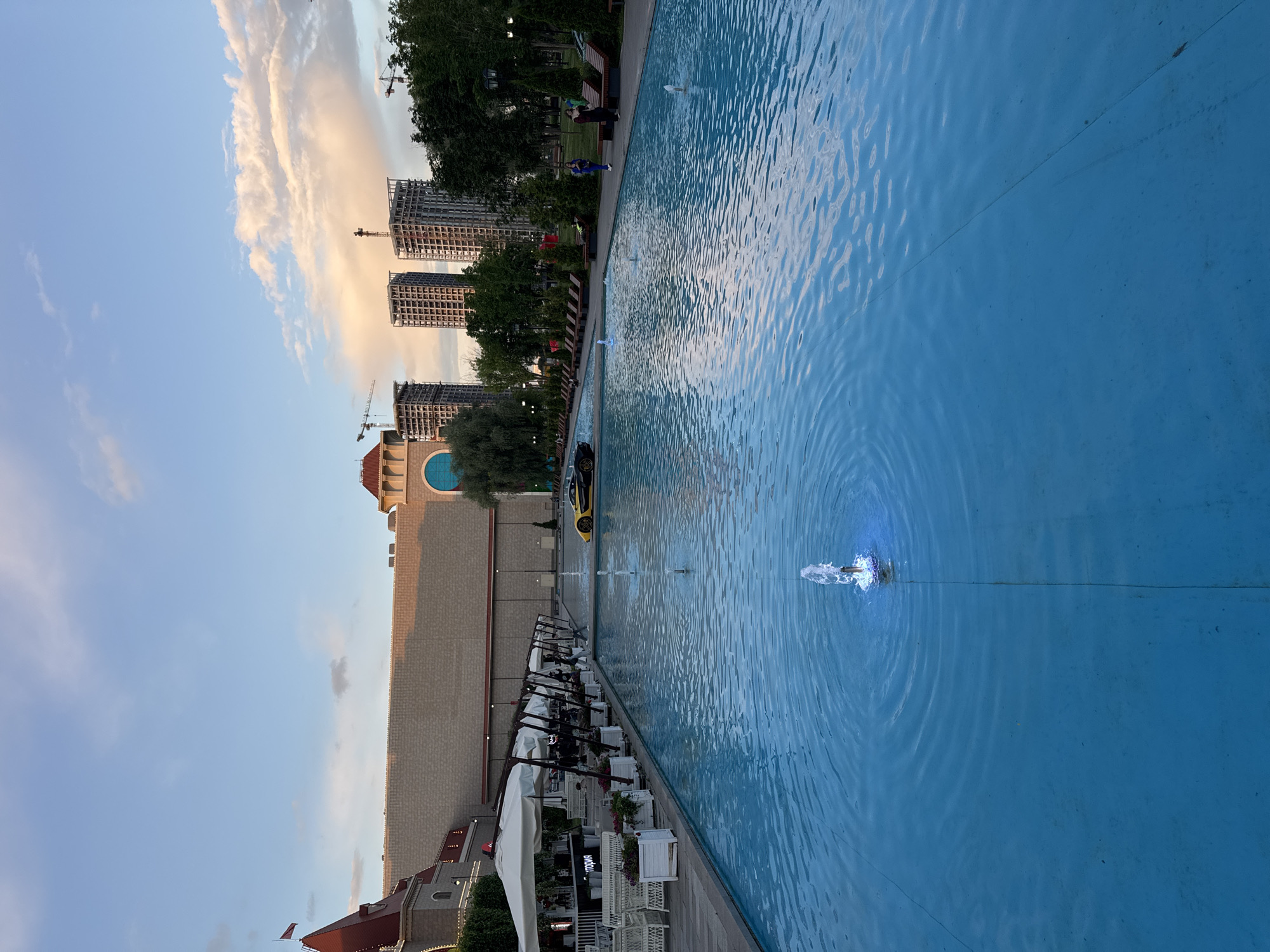 The image depicts a public outdoor area in Moscow, Russia, featuring a large, rectangular blue swimming pool with several small water fountains. In the background, there is a building with a red roof and light-colored brick walls, reminiscent of a historical or entertainment complex. Adjacent to this structure are several tall, unfinished buildings under construction, with visible scaffolding and cranes, indicating urban development. Trees and park benches are also present, suggesting a recreational space. A yellow sports car is parked by the edge of the pool. Several people are visible in the scene; some are seated on lounge chairs, while others are walking. The sky is partly cloudy with hues of blue and pink, suggesting either sunrise or sunset.