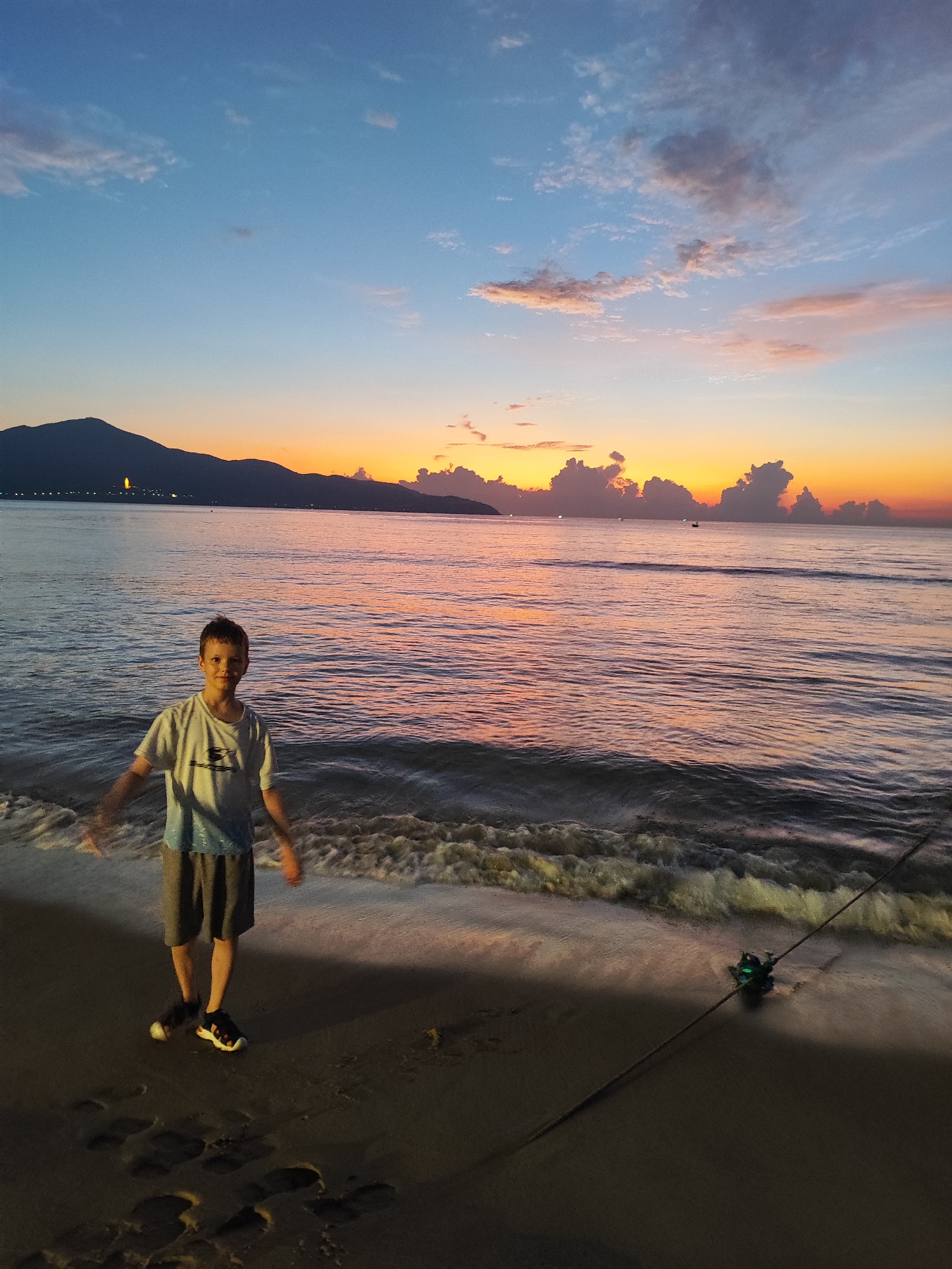A medium shot captures a young boy standing on a beach with the ocean waves lapping at his feet, set against the backdrop of a vibrant sunset. The boy is wearing a light-colored T-shirt and grey shorts, with athletic shoes. His arms are slightly outstretched at his sides. To his right, a fishing rod rests on the sand, partially submerged in the water.
The ocean reflects the colors of the sunset, creating a blend of oranges, pinks, and purples. In the distance, a dark mountain silhouette is visible, punctuated by a few lights. The sky transitions from a light blue at the top to the warm colors of the sunset near the horizon, with clouds adding texture to the sky.
The location is the beach in Đà Nẵng, Vietnam, as suggested by the mountain range that's part of the local scenery. The photo was likely taken during the golden hour, just before sunset, given the position of the sun and the lighting conditions.