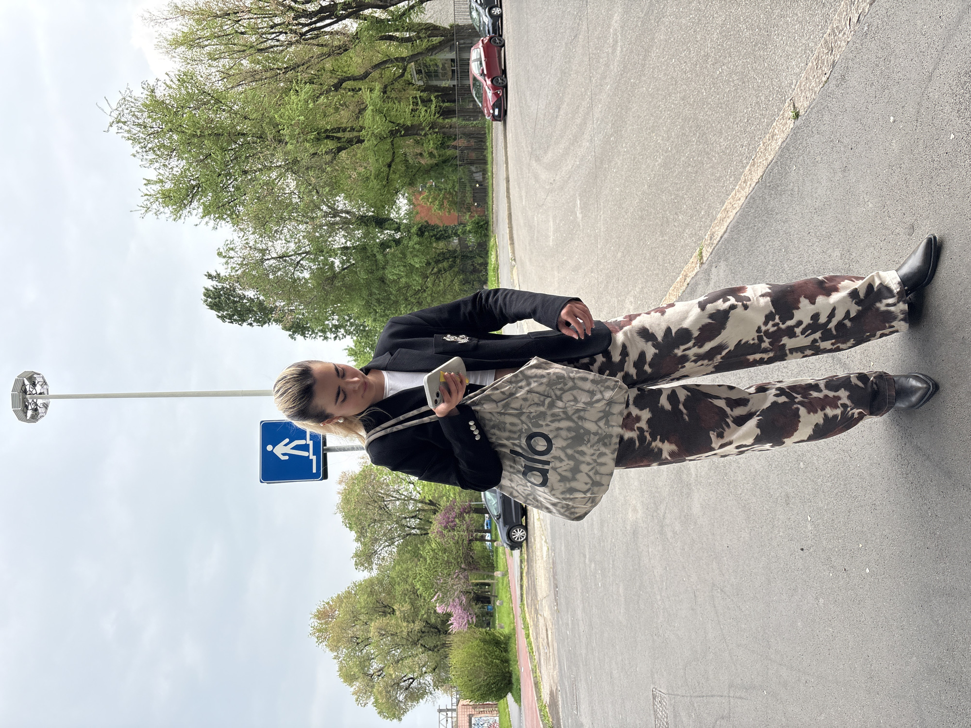 A woman stands on a city street in Milan, Italy. She is wearing a black blazer, white top, cow-print pants, and black shoes. She is holding a smartphone in her right hand and an Alo brand bag hangs from her left shoulder. Behind her is a sign indicating the direction to the stairs. The street is lined with trees and parked cars, and the sky is overcast.