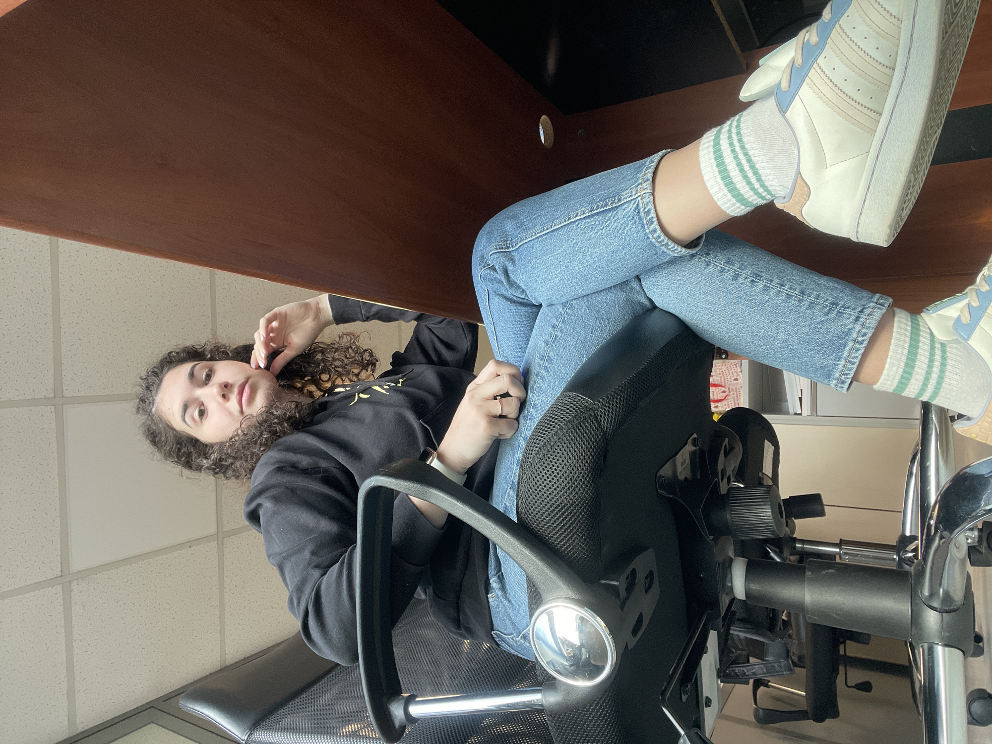 A young woman with curly hair is sitting in an office chair, her legs crossed on a desk. She is wearing jeans, a black hoodie, and white sneakers with light blue accents and green-striped socks. She is looking at the camera, and her right hand is near her face. The office has a light-colored tile ceiling and a brown desk. The location cannot be determined to be in the city of Sofia, Bulgaria from the image alone.