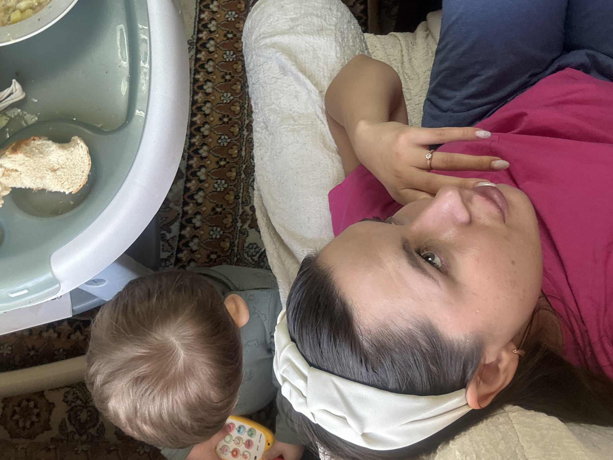 The image shows a person, possibly a parent, wearing a pink shirt, dark blue jeans, and a white headband. They are lying on a light-colored surface, possibly a couch or bed. A child with light brown hair is near them, holding a yellow toy with colorful buttons. To the left of the child is a highchair tray with food, including a piece of bread and a plate with some food items. The floor is covered with a patterned rug. The location is Красный Маяк, Russia.