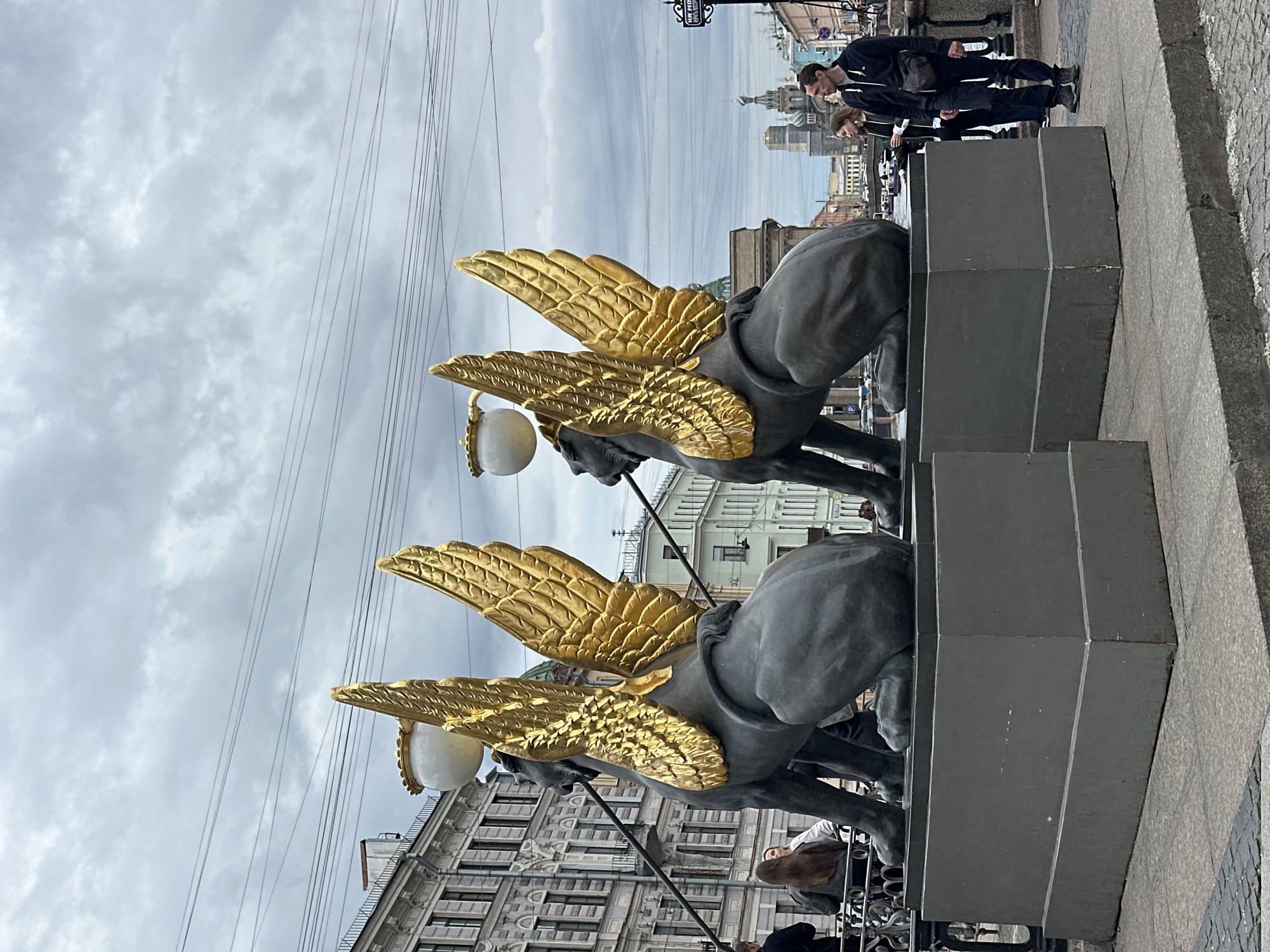 The image captures a view of the Sphinx Bridge in Saint Petersburg, Russia. In the foreground, two dark-colored sphinx statues with large, golden wings are prominently displayed on pedestals. They face away from the viewer, with the statue on the left slightly closer. Above the sphinxes, lamps with white spherical shades are mounted.
In the background, a cloudy sky with visible power lines stretches across the frame. To the left, a building with multiple stories, arched windows, and decorative architectural elements is visible. Further in the background, other city buildings and structures can be seen, including church domes.
Several people are present in the scene. On the far left, a person wearing a dark jacket is partially visible. Near the left sphinx, two individuals, one with long brown hair, are standing. To the right of the sphinxes, a man in a dark hoodie and black pants is walking, and a woman in a dark jacket is beside him. Other people are dispersed further in the background. The scene appears to be an outdoor public space, likely a bridge or embankment.