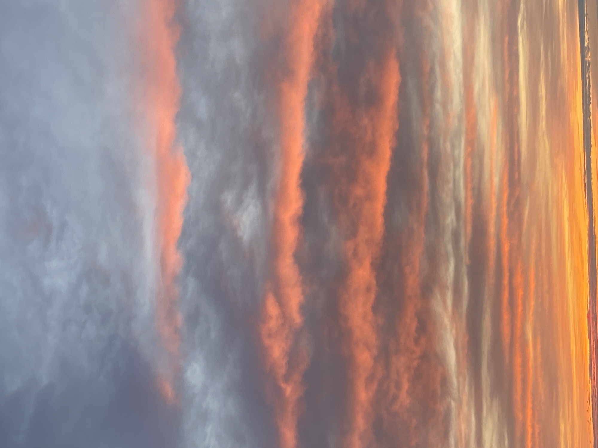 The image shows a vertical view of the sky during sunrise or sunset. The clouds display a striated pattern, with streaks of orange and reddish hues intermingling with grayish-blue tones. The right side of the image shows a gradient of colors ranging from a pale yellow to a deep orange. No people or objects besides the clouds are visible. Location information is unavailable.