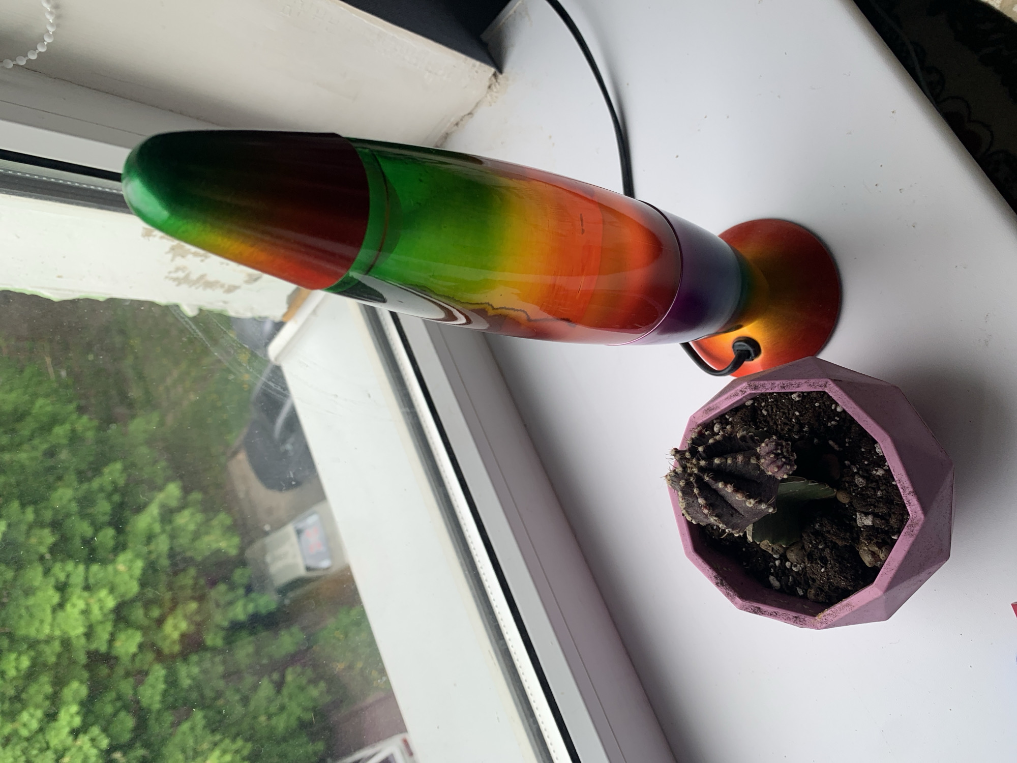 On a white windowsill in Unknown, Unknown, is a lava lamp horizontally oriented next to a potted cactus. The lava lamp is multicolored, transitioning from red and green on one end to orange and purple on the other. A black electrical cord extends from the lamp's base. The cactus is in a geometric, light purple pot. Outside the window, dense green foliage is visible.