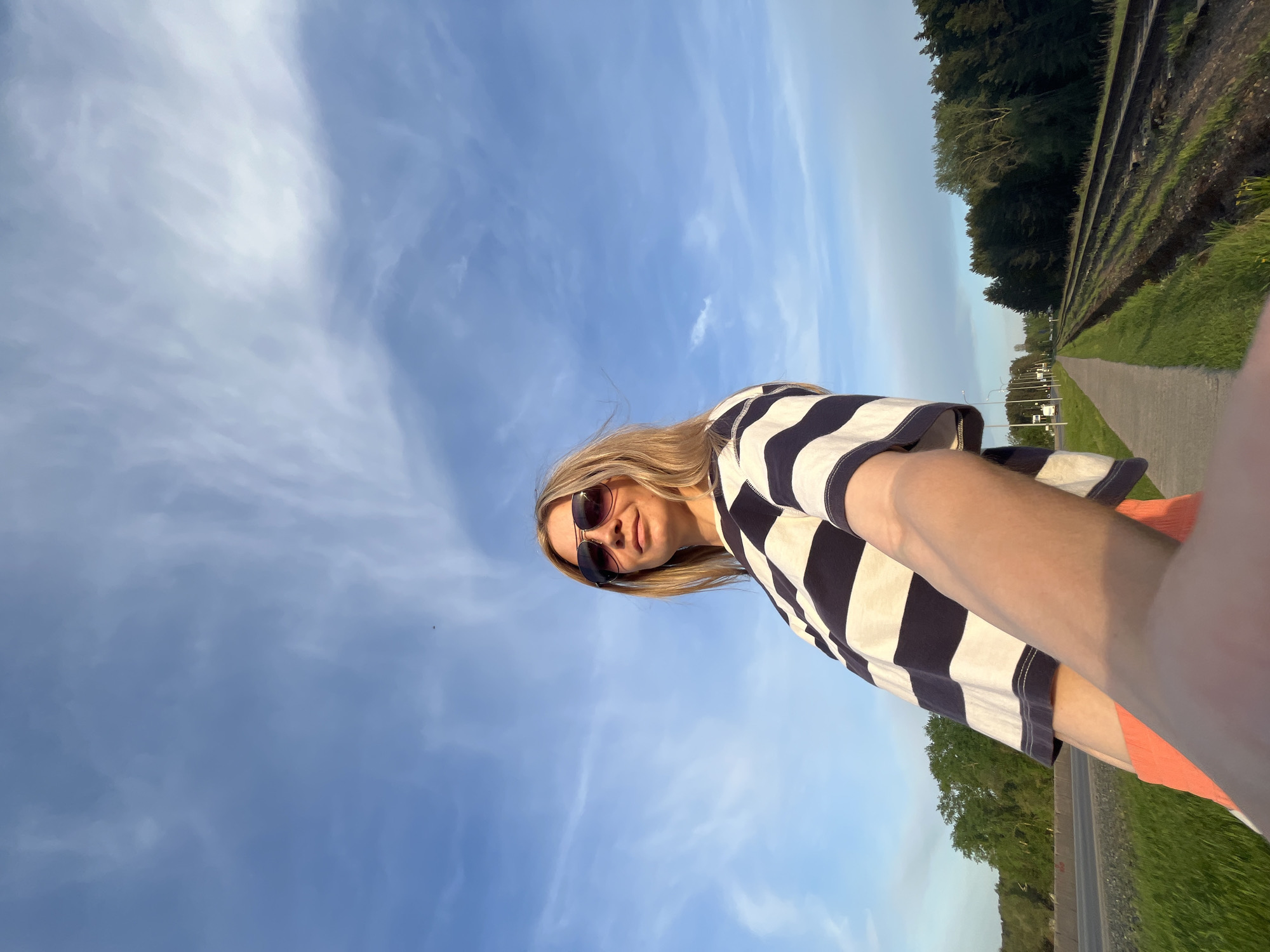 A woman stands on a path next to train tracks on a clear day, holding her phone up and taking a selfie. She has blonde hair, sunglasses, and wears a striped shirt. The sky is blue with light clouds. In the background are trees, tracks, a path, and possibly street lights. It is unclear where this image was taken within the city of Chaikovsky, Russia.