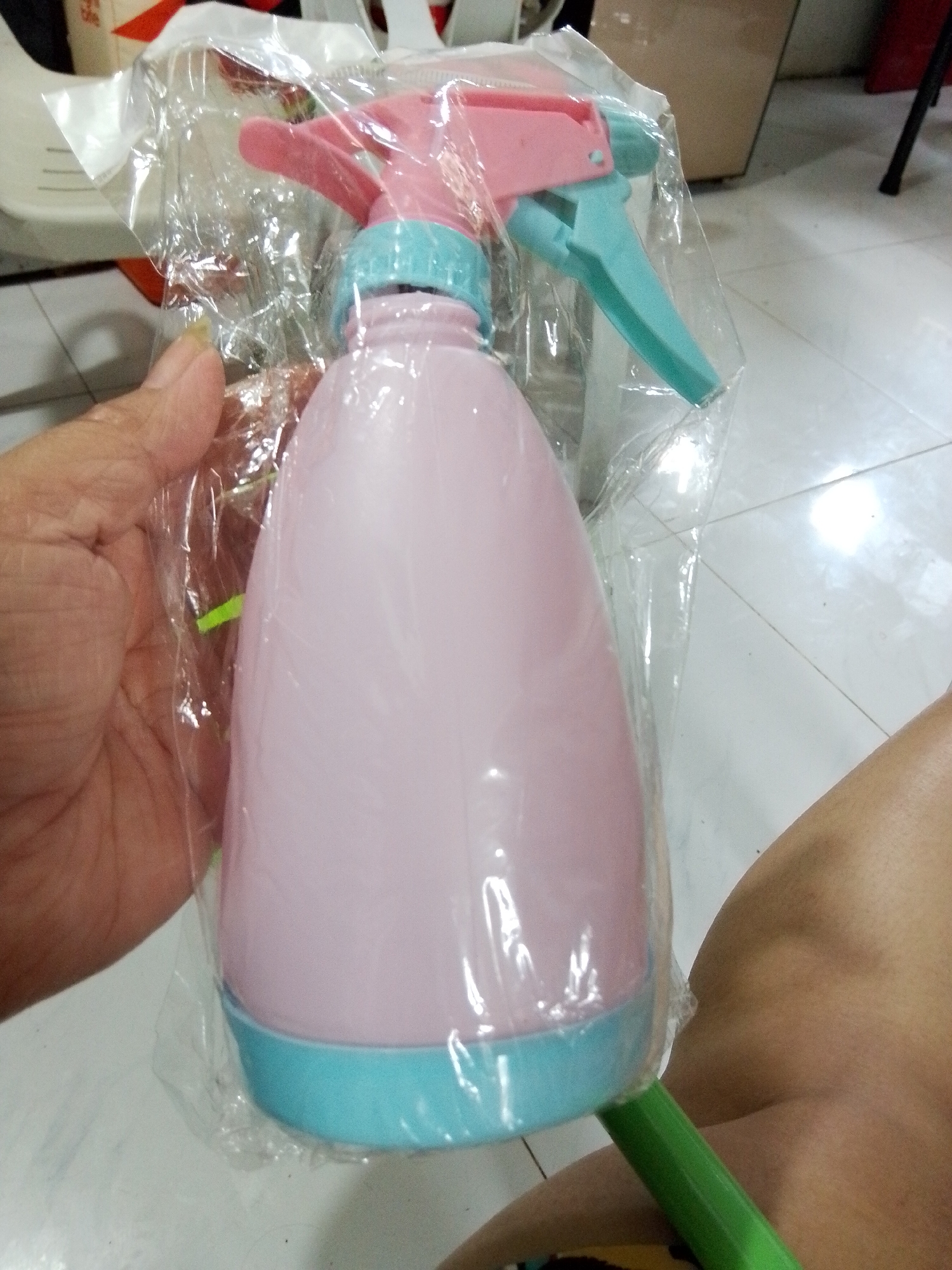 A close-up indoor shot captures a person's hand holding a new, pastel-colored spray bottle encased in clear plastic packaging. The spray bottle itself is primarily light pink, with a distinctive pastel turquoise band at its base and matching turquoise elements on the spray nozzle's collar and trigger handle. The main spray head is pink, complementing the bottle's body.
The person's hand, positioned on the left, holds the packaged item firmly. Their fingernail is visible and has a slightly yellowish tint. In the background, a light-colored tiled floor is prominent, suggesting an indoor setting, likely a home or shop in Davao City, Philippines. Other background elements are blurred, hinting at furniture or storage items, with a dark, possibly metallic, object in the upper right. A vibrant green object, possibly a handle or part of another item, peeks into the frame from the bottom right. The lighting is consistent with ambient indoor conditions, and no specific time of day or emotional state can be discerned. There is no visible text on the spray bottle or its packaging.