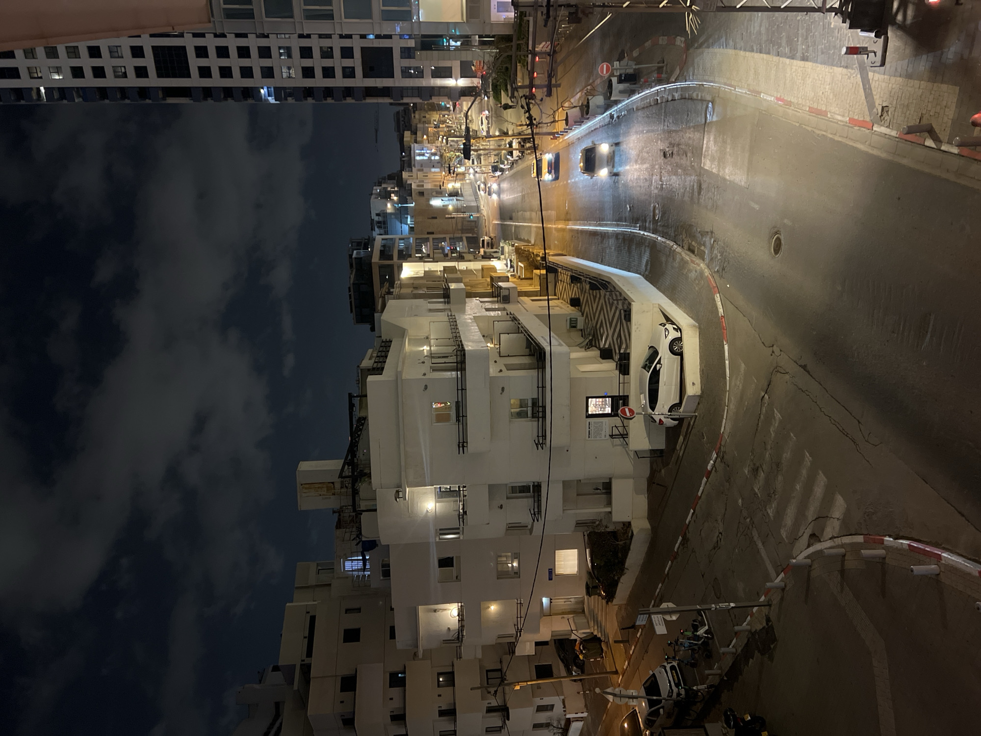 This is an elevated nighttime view of a street scene in Tel Aviv, Israel. In the background, a street stretches into the distance, lit by streetlights. Cars with illuminated headlights drive along the street. To the left, multiple residential buildings are visible, their facades illuminated by interior lights. The sky is dark, with scattered clouds. In the foreground, a curb marked with red and white paint, and a section of the road with a pedestrian crossing. Several parked vehicles can be seen near a white building.