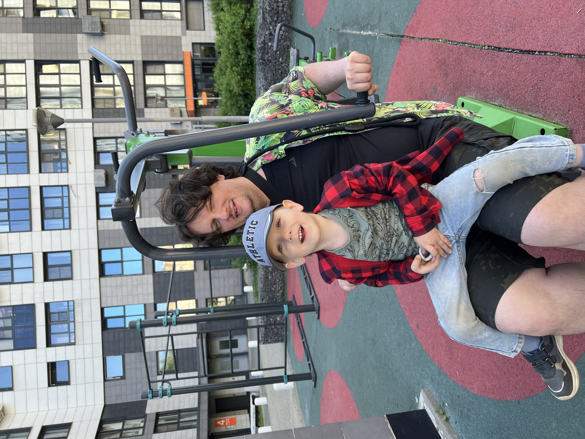 A man and a young boy are captured in a medium close-up, outdoors, on what appears to be a playground or outdoor gym area, with residential buildings in the background. The image is rotated 90 degrees clockwise, placing the subjects horizontally across the frame.
The man, positioned on the left, has dark, curly hair and is looking towards the viewer with a slight smile. He is wearing a black t-shirt and a green short-sleeved shirt with a tropical pattern. His left arm is bent, and his hand is gripping a handle of a piece of exercise equipment, which is green and grey.
Sitting on the man's lap or close to him is a young boy, also smiling broadly at the camera. He wears a light blue baseball cap with "ATHLETIC" written on it in white letters, a green t-shirt with a patterned design, and a red and black plaid long-sleeved shirt. He is wearing light blue ripped jeans and black sneakers with white soles. The boy's hands are clasped together, and he appears to be holding a small white object.
The setting includes a red and green padded ground surface, characteristic of playgrounds. In the background, modern apartment buildings with grey, white, and black facades and numerous windows are visible. Some windows reflect the sky or surrounding environment. Green foliage, likely trees or bushes, can also be seen in the mid-ground. Another piece of metal gym equipment, possibly a monkey bar or pull-up station, is partially visible in the background on the right. The lighting suggests it is daytime. The general atmosphere is casual and happy.