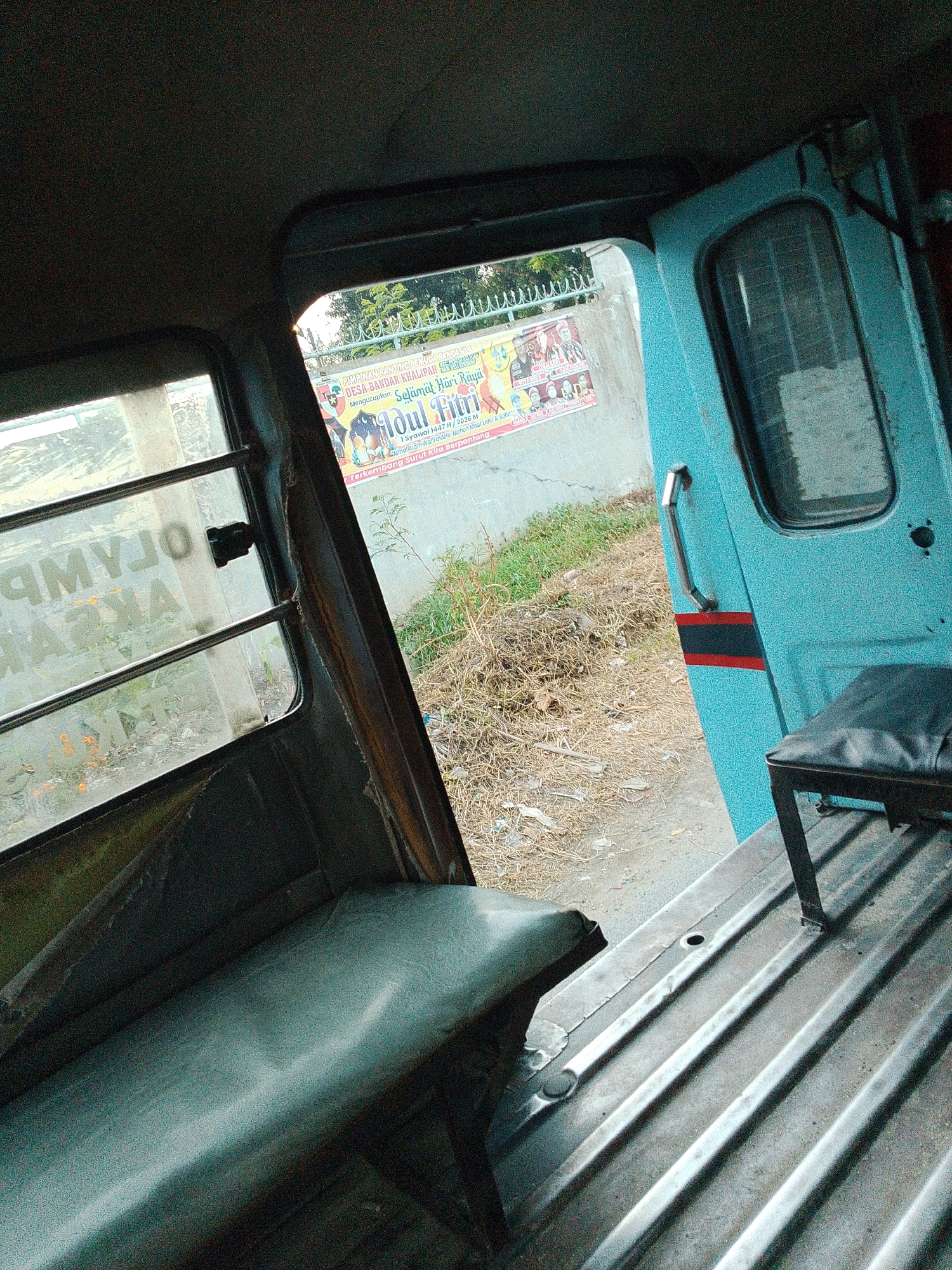 The image depicts the interior of a vehicle, likely a type of public transport, viewed from a low angle. To the left, a worn, teal-colored bench seat runs along the side. Above it, a window is visible with horizontal bars and text that appears to be in reverse, possibly "OLYMPUS AKAR." The interior of the vehicle has a dark, textured ceiling.
The door on the right side of the vehicle is open, revealing the exterior. The door is painted a vibrant teal with a thin red and black stripe. A chrome handle is attached to the door. Outside, a concrete wall is visible with a poster attached to it. The poster features text in Indonesian, including "PIMPINAN RANTING Muhammadiyah DESSA BANDAR KHALIPAH" and "Selamat Hari Raya Idul Fitri." This indicates the location is in Bandar Khalipah, Indonesia. Beyond the wall, dry grass and some vegetation are present, along with a black fence and trees.
In the foreground, the floor of the vehicle is visible, made of corrugated metal with a dark, ribbed texture. A small, dark seat or bench is positioned to the right, with a black upholstered seat on a metal frame. The overall lighting is dim, with some natural light entering from the open door and window. The image has a grainy texture.