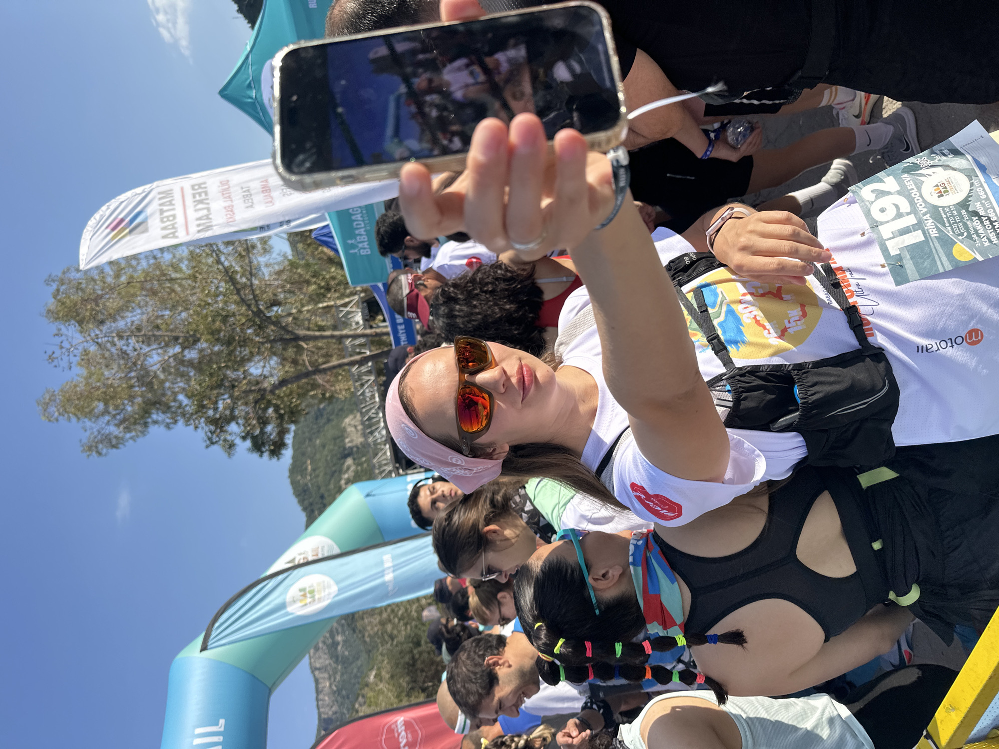 A medium eye-level outdoor shot depicts a woman taking a selfie with her mobile phone in the city of Fethiye, Turkey. The city is home to the Babadag Ultramarathon.
The woman is wearing a white shirt, a pink bandana, and orange sunglasses. She is surrounded by other people, some of whom are also wearing running attire.
The shot includes an archway decorated with blue and white balloons. Banners with Turkish text are visible in the background. Another sign appears with the name "IRINA VODOLEEVA".