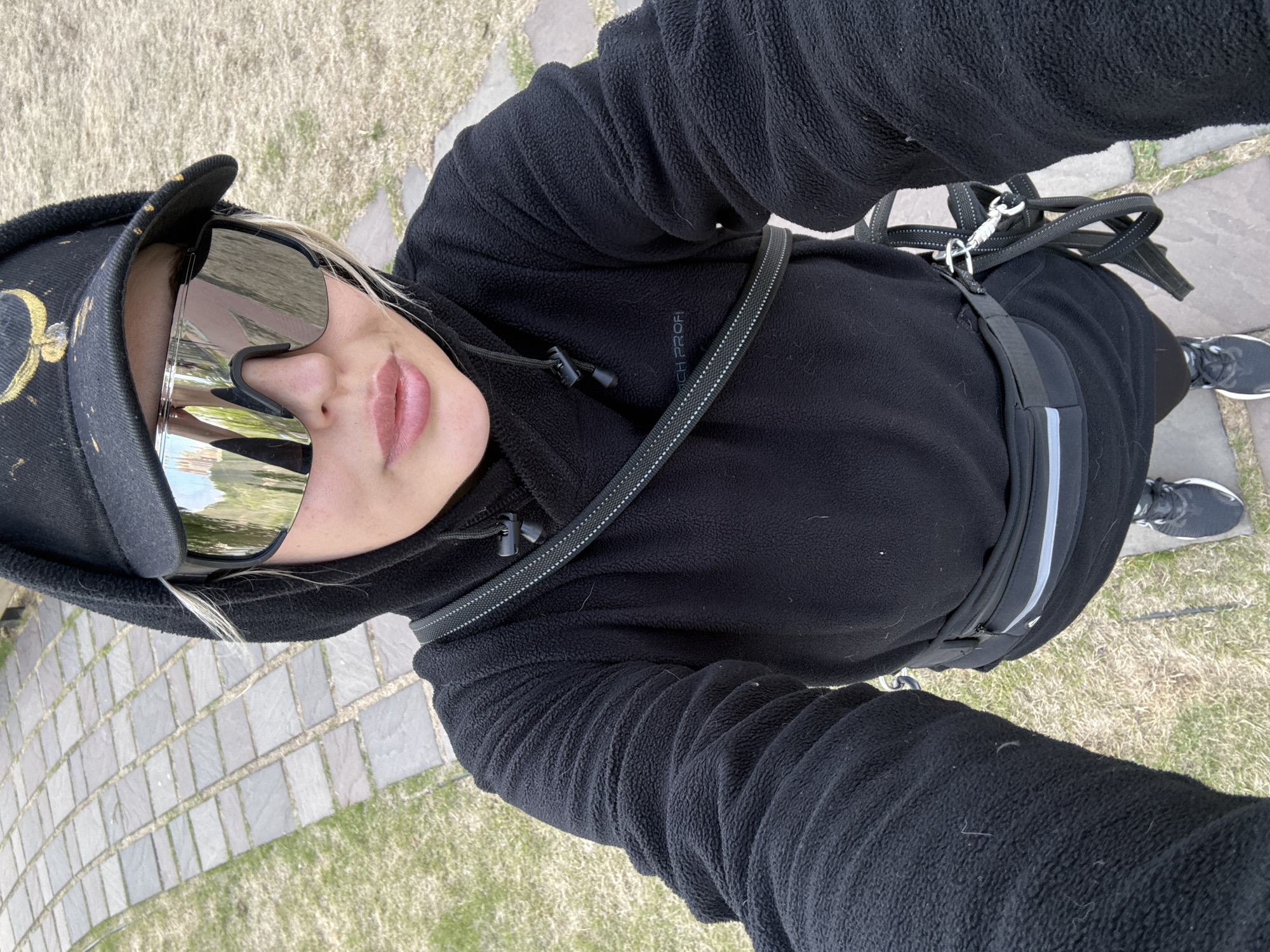 A person wearing a black fleece jacket, a black baseball cap with a yellow design, and mirrored sunglasses is looking directly at the camera. The person is standing on a paved walkway consisting of grey rectangular stones with grass growing between them. A black dog leash is slung across the person's chest, and a black running belt with reflective stripes is around their waist. The person is wearing black athletic shoes with white soles. The ground is covered in dry grass. The photograph appears to have been taken at a high angle. The location is Krasnoyarsk, Russia.