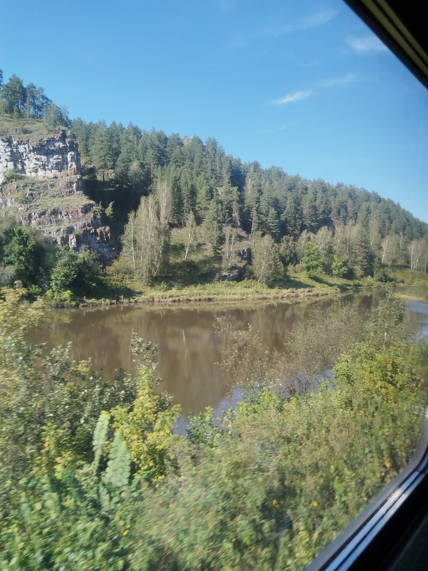 The image shows a natural landscape with a river, trees, and a rocky cliff. The river is in the foreground, reflecting the sky and surrounding trees. The left side of the image features a steep, rocky cliff face with vegetation growing on its slopes. The top of the cliff is covered with a dense forest of coniferous and deciduous trees. The sky is clear and blue with a few wispy clouds. The scene appears to be a scenic view from a moving vehicle, likely a train, as suggested by the curved window frame visible on the right side of the image. The image location is identified as Vyazovaya, Russia.
