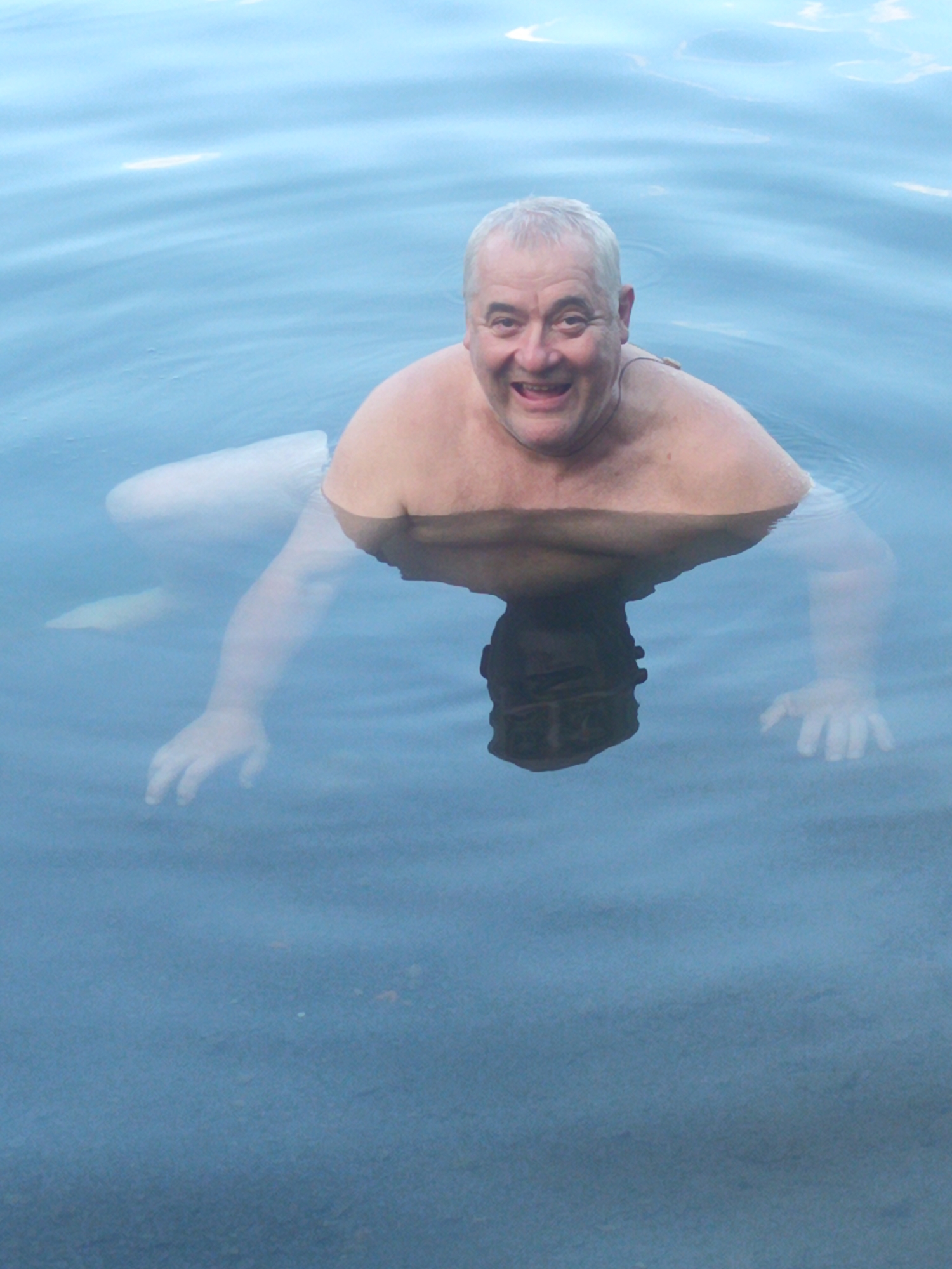 A man is swimming in a body of water, likely a river or lake, with his upper body exposed. The water is calm with light ripples on the surface. The man is smiling at the camera, and his head and shoulders are clearly visible. His arms and torso are submerged or partially submerged. His reflection is visible in the water, showing the inverted image of his head and upper body. The environment appears to be an outdoor setting, with the water reflecting the sky. The specific location in Uzhhorod, Ukraine is not determinable from the image alone, as it lacks identifying landmarks.