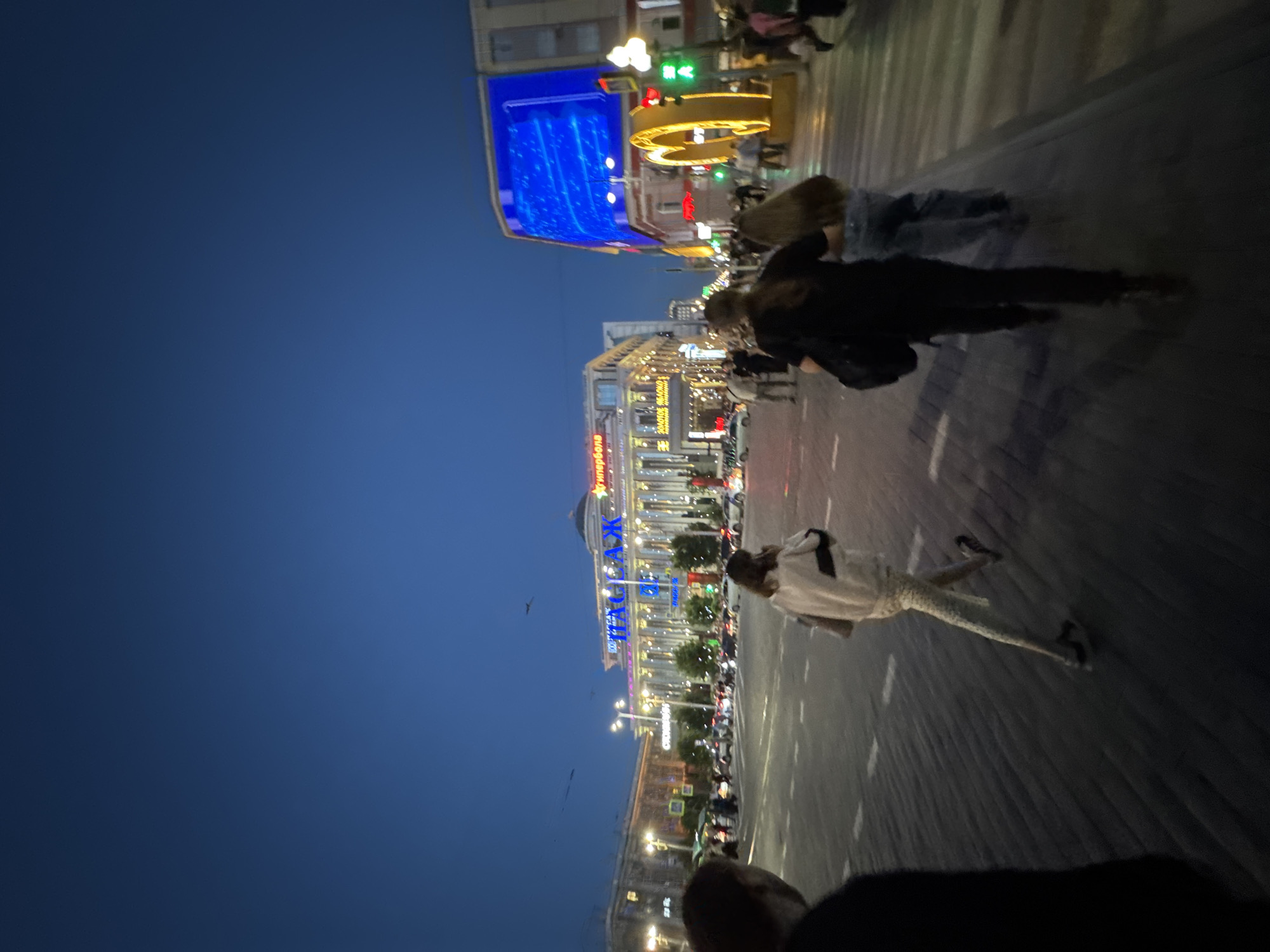 This image captures a street scene at dusk in Yekaterinburg, Russia, specifically on Yekaterinburg's main square, 1905 Square. A large building with illuminated signage in Russian, likely a commercial establishment, is visible in the background. The architecture features classical elements with columns. To the right, a large LED screen displays a blue graphic, and a prominent yellow arch structure is illuminated. Several people are walking across a paved street, which is marked with pedestrian crossing lines. The sky is a deep twilight blue. Vehicles are present on the street, and traffic lights are visible, indicating a busy urban intersection. The overall atmosphere suggests a lively evening in a city center.