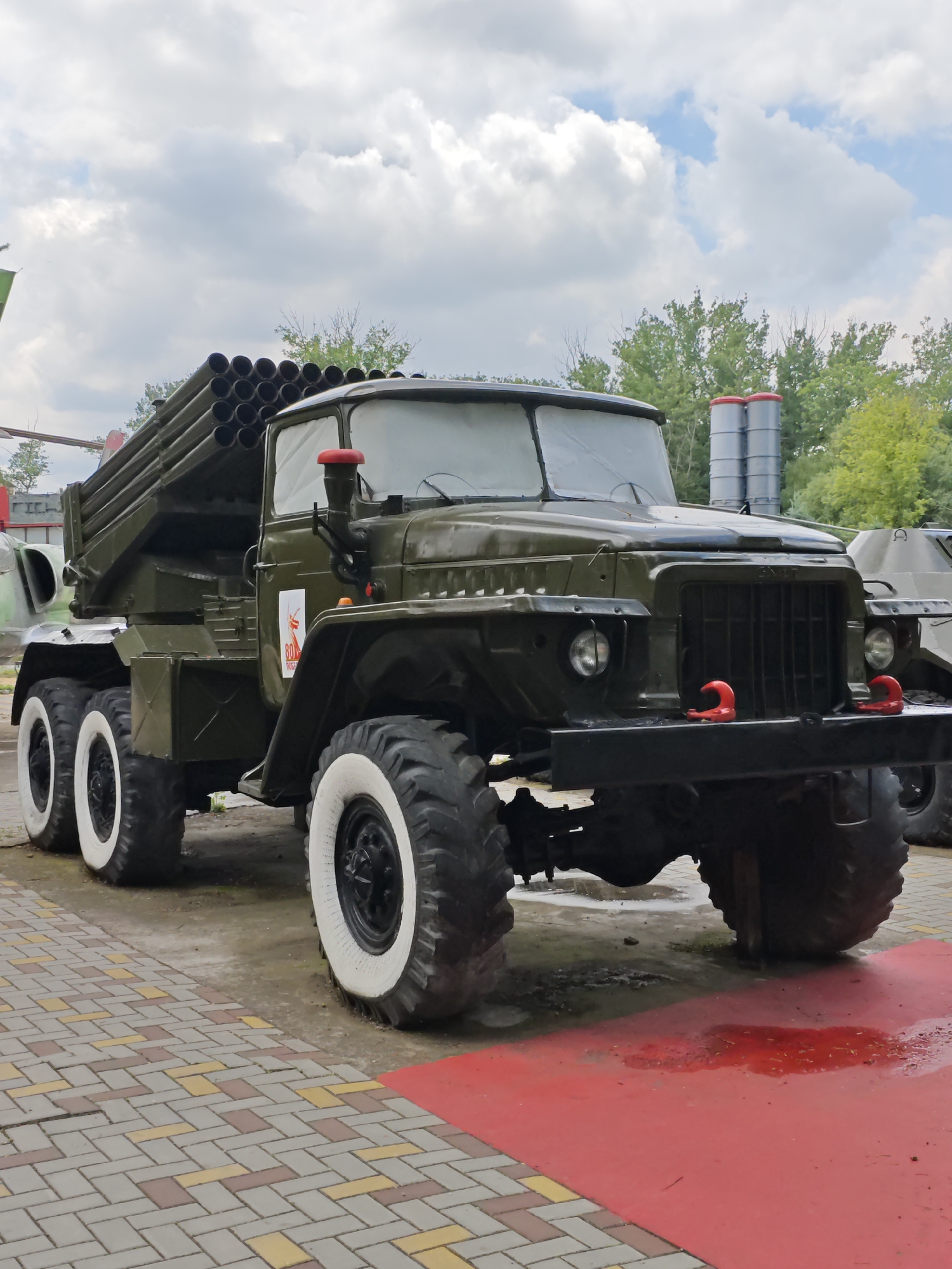 A military vehicle, identified as a BM-21 Grad multiple rocket launcher, is parked on a paved surface with a red painted area in front of it. The vehicle is dark green and has a large number of tubes mounted on its rear, which are part of the rocket launching system. It has large, rugged tires with white sidewalls. The scene appears to be an outdoor exhibit or display, possibly at a museum or memorial, given the presence of other military hardware in the background, including what appears to be a missile system and part of another vehicle. The sky is partly cloudy, with a mix of white clouds and blue sky. The ground in front of the vehicle is wet, with some red liquid pooling on the red painted surface, suggesting recent rain or a spill. The location is the city of Cherkessk, Russia.