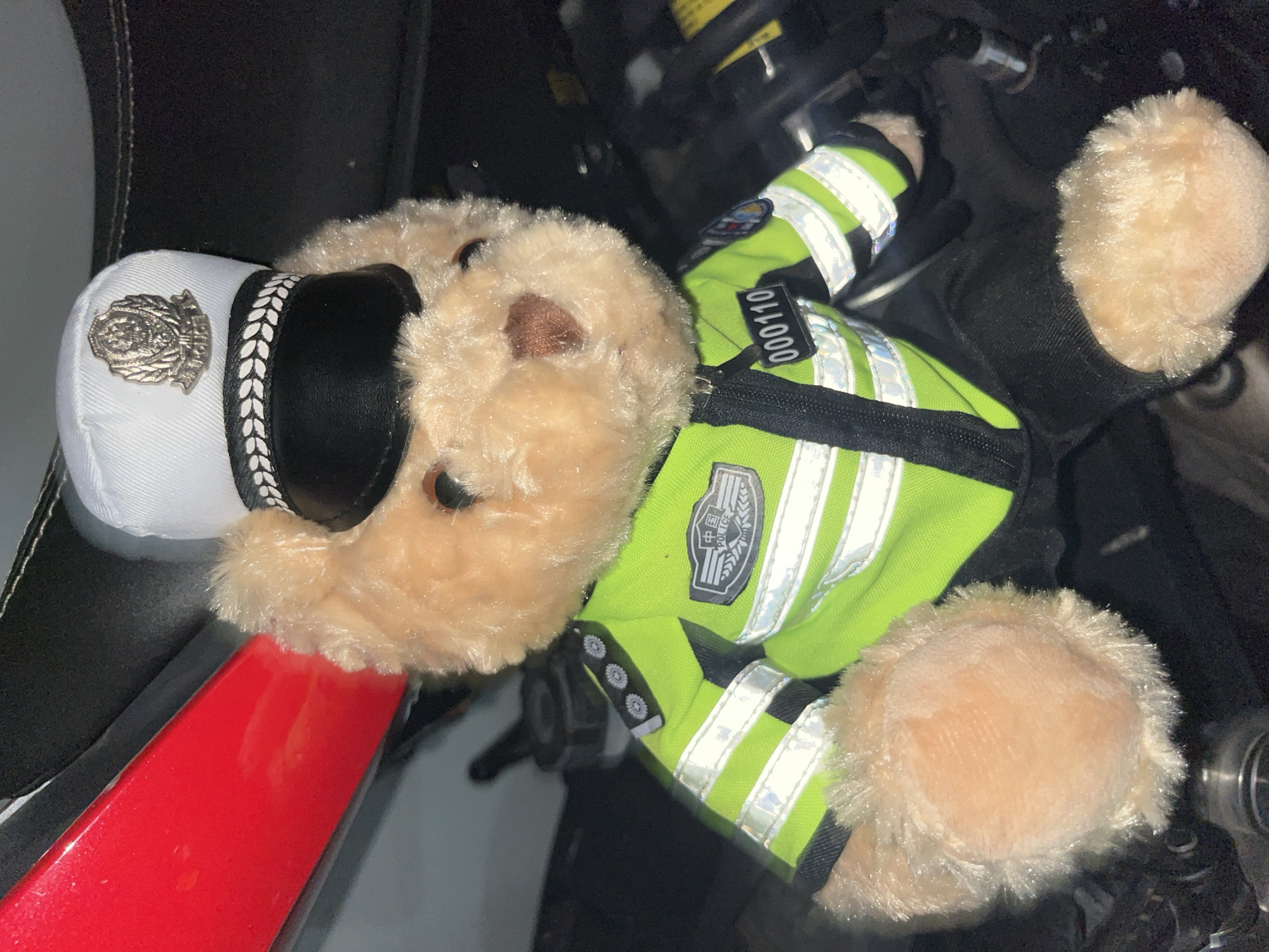 A plush teddy bear wearing a police uniform is the central subject of this image. The bear is positioned in a dark environment, with a hint of a red object to its left, possibly part of a vehicle. The bear's police uniform includes a white cap with a silver emblem and a black brim, a bright green high-visibility jacket with reflective stripes, and black pants. The jacket has badges and a number tag that reads "000110". The background appears to be a mechanical or engine compartment, with various dark-colored components and wires visible. The lighting is dim, creating shadows and highlighting the reflective elements of the uniform. The overall impression is that the teddy bear is posed within a vehicle or a similar setting, likely in Yuzhong District, China, given the presence of Chinese characters on the badge and the context provided.