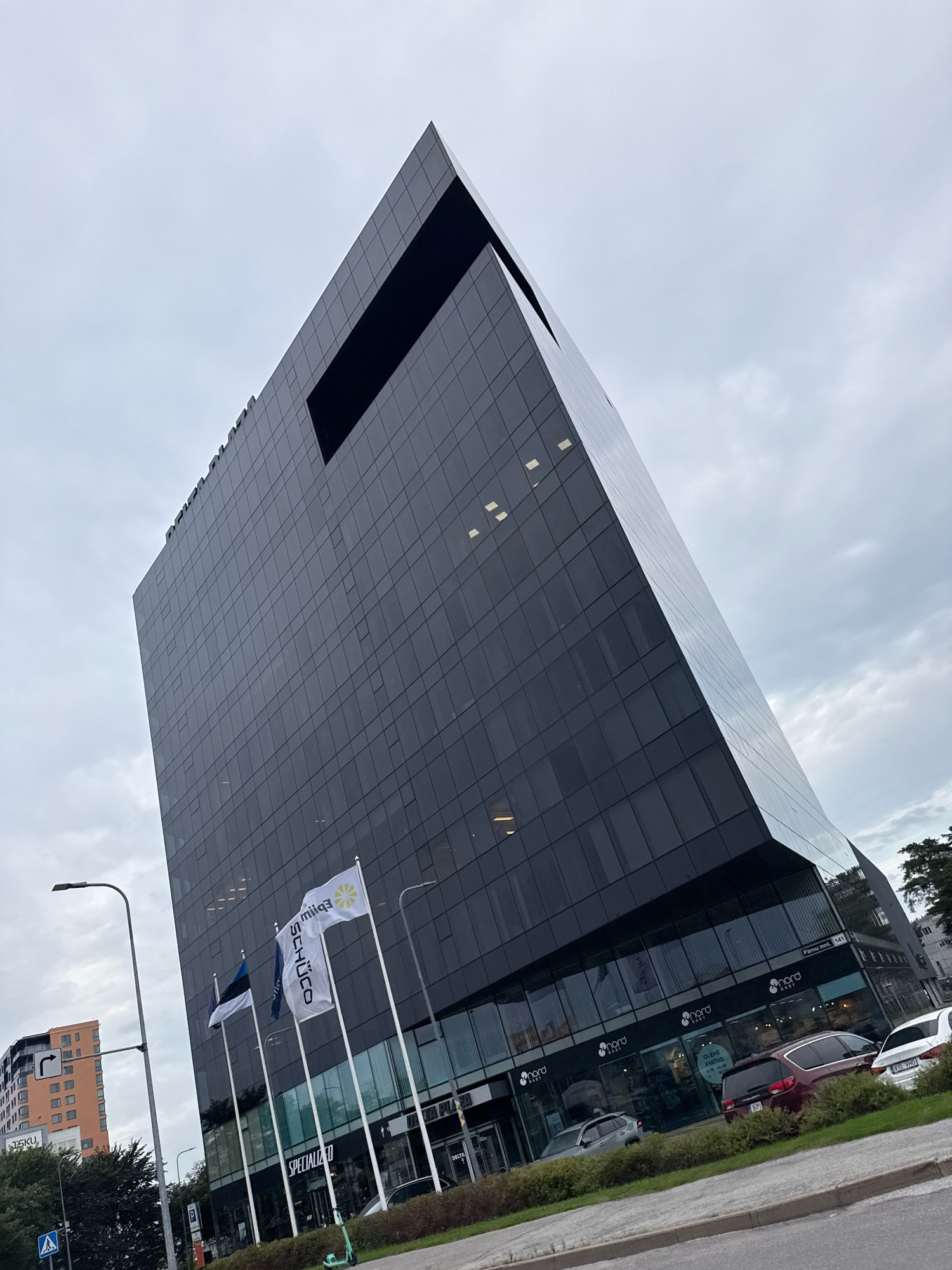 The image shows a modern, angular, dark glass-clad building in Tallinn, Estonia, on a cloudy day. The building features a large, dark rectangular cutout near the top and a slanted facade. Several flags are displayed outside the building, including the Estonian flag and flags with white fabric and logos. The ground floor of the building has large glass windows with signage for "NORD BABY" and "SPECIALIZED". A street with parked cars and a traffic light is visible in front of the building. In the background, another apartment building with orange facade is seen. The scene depicts an urban environment with commercial and residential elements.