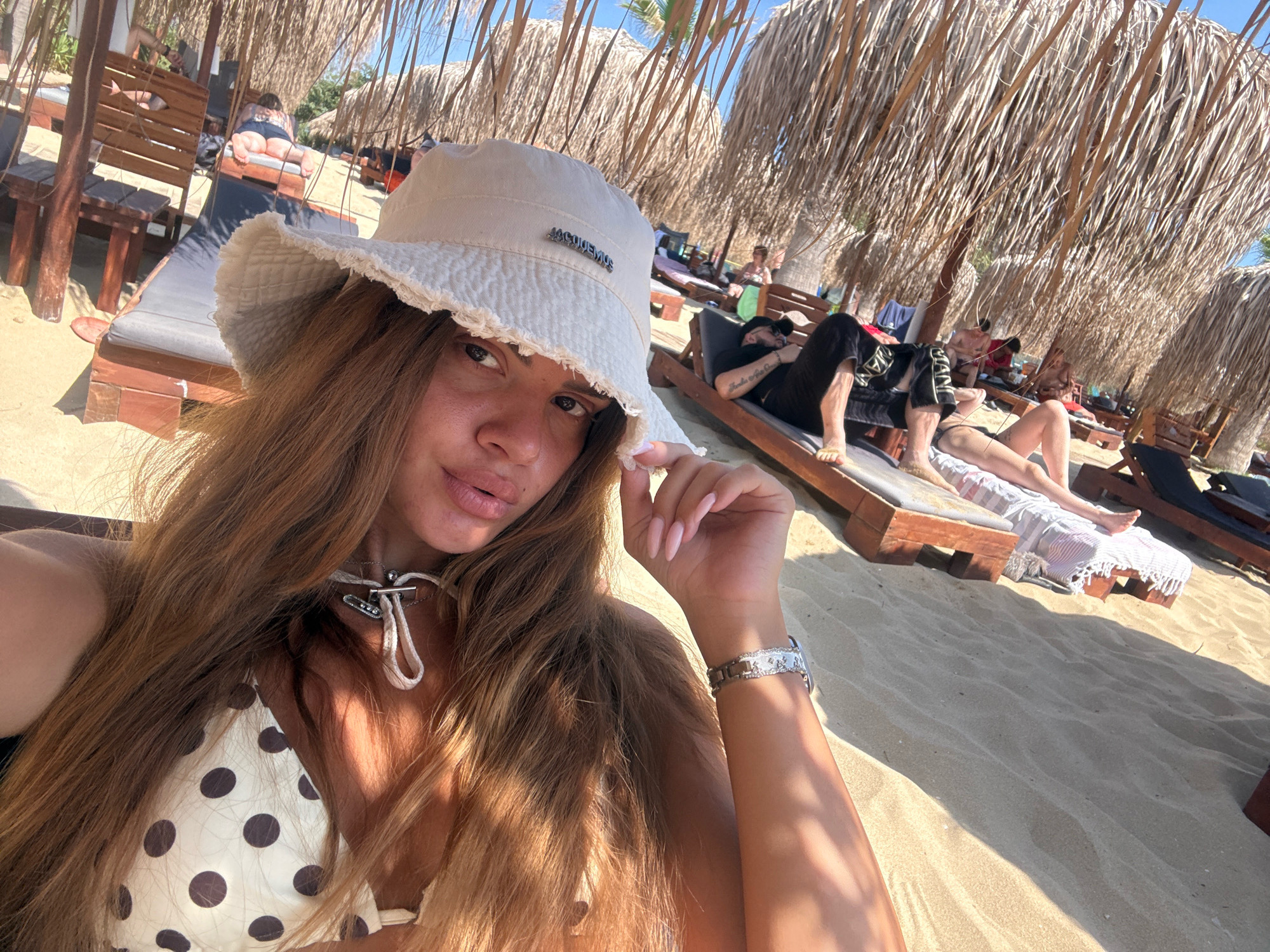 The image shows a woman in a white bucket hat and a polka dot bikini top taking a selfie on a sandy beach in Nea Peramos, Greece. She is wearing a necklace and a watch. In the background, several people are relaxing on lounge chairs under thatched umbrellas. The sun is shining, casting shadows on the sand. The scene appears to be a beach resort or a public beach with amenities.