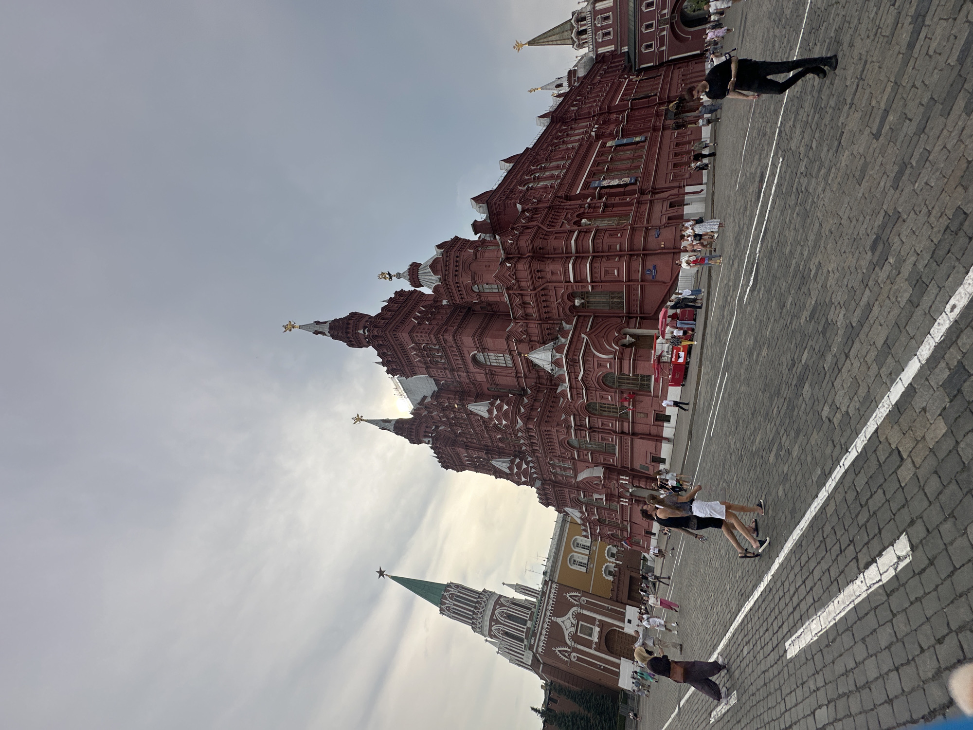 This is a wide-angle, low-angle, outdoor, daylight photograph of Red Square in Moscow, Russia. The iconic State Historical Museum, a large red brick building with ornate architecture and numerous spires, dominates the right side of the frame. To the left, a portion of the Spasskaya Tower of the Moscow Kremlin is visible, distinguished by its green spire topped with a red star. The ground is paved with cobblestones, with white lines indicating pedestrian paths. Numerous people are walking across the square in various directions. Some are dressed casually in t-shirts, shorts, and dresses. Two food carts are visible near the museum. The sky is overcast with light clouds.