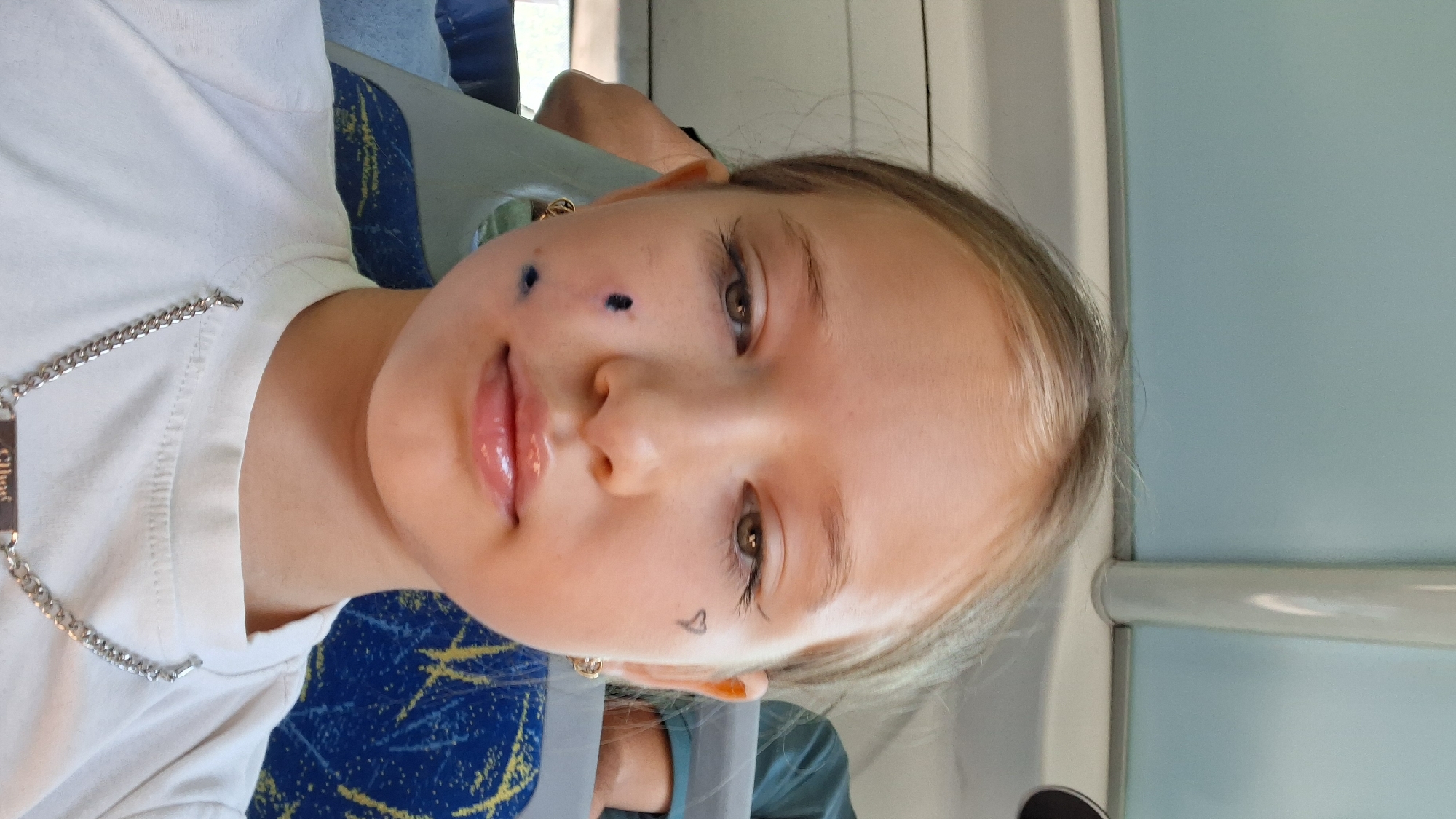 A young girl with blonde hair pulled back is looking into the camera. She is wearing a white t-shirt and a silver chain necklace. On her left cheek are three dark marks, possibly ink stains or temporary tattoos. A small heart is drawn with eyeliner on her right cheek. She is seated on a bus, with a blue and yellow patterned seat visible behind her. The background is blurred, but appears to be the interior of a public transport vehicle.