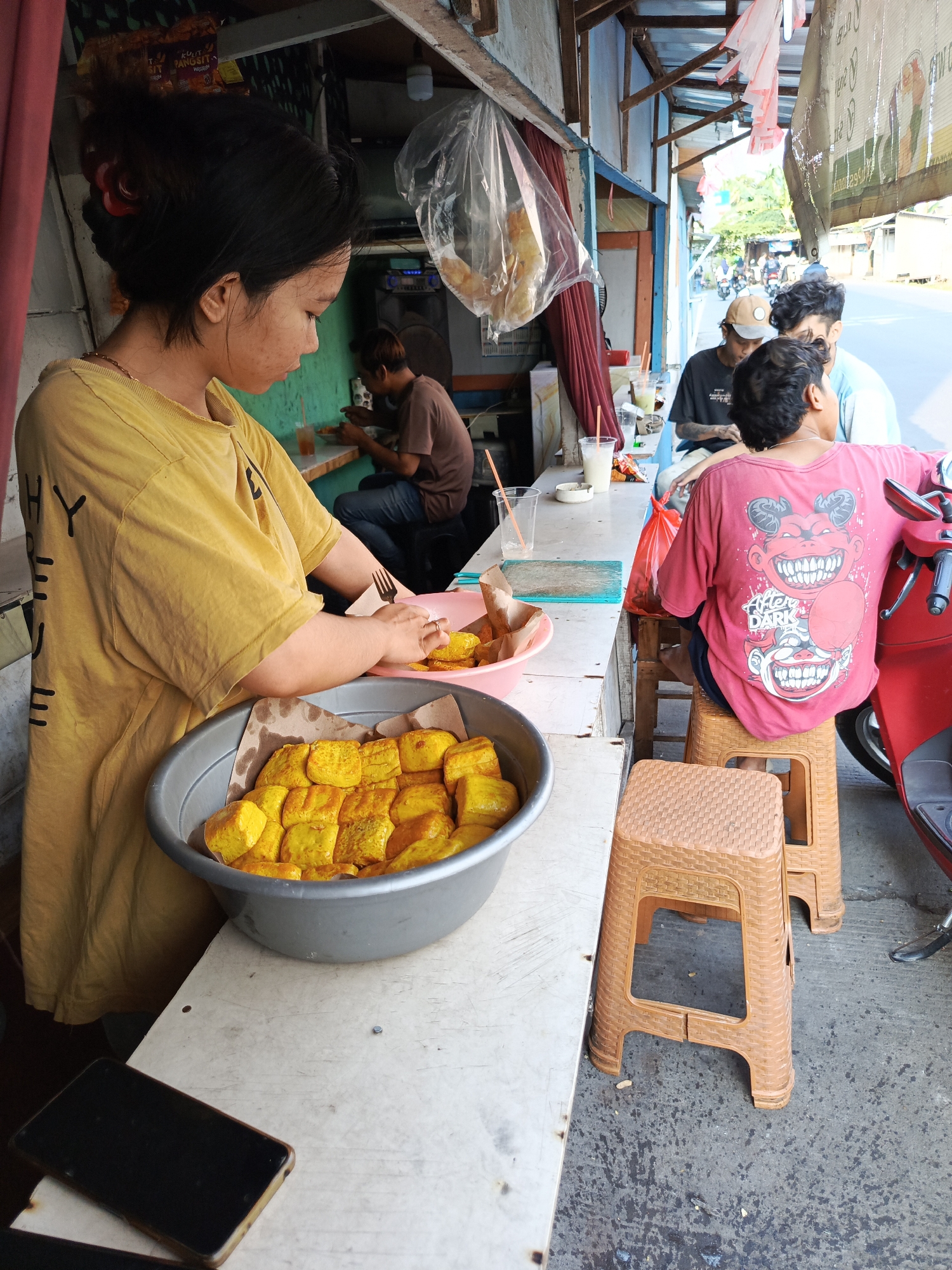 The image depicts a street food stall in Tangerang, Indonesia, likely on a bright, sunny day based on the lighting. In the foreground, a young woman wearing a yellow t-shirt is preparing food. She is standing behind a counter that displays a large metal bowl filled with golden-brown fried tofu. To her left, a dark-colored mobile phone rests on the counter. Behind her, a man in a brown shirt is seated and eating, and further back, two other young men are also seated. One of them is wearing a pink t-shirt with a graphic design. A red motorcycle is parked to the right of the frame, partially obstructing a view of the street which appears to be a main road with passing vehicles. The stall itself is constructed with a wooden frame and a corrugated metal roof. Plastic bags and various food items are visible hanging or placed around the stall, suggesting it is an active place of business. The overall scene conveys a typical urban street food environment in Indonesia.