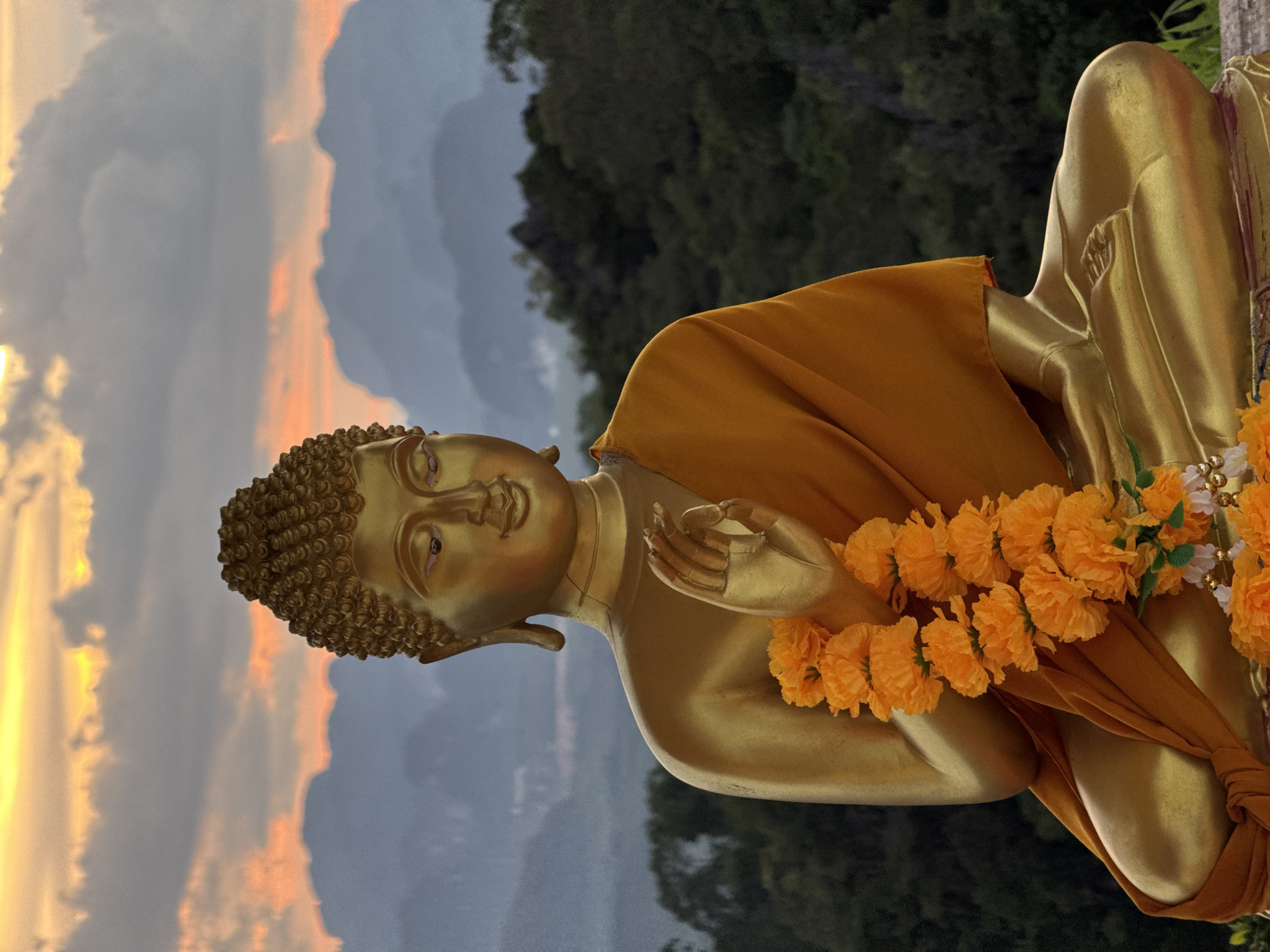 A gold-colored statue of Buddha is seated in a lotus position, adorned with an orange flower garland. The statue is positioned outdoors, with a lush green forest and mist-covered mountains in the background. The sky above displays a dramatic sunset with clouds illuminated by orange and yellow light. The location is identified as Ban Khao Tang, Thailand.