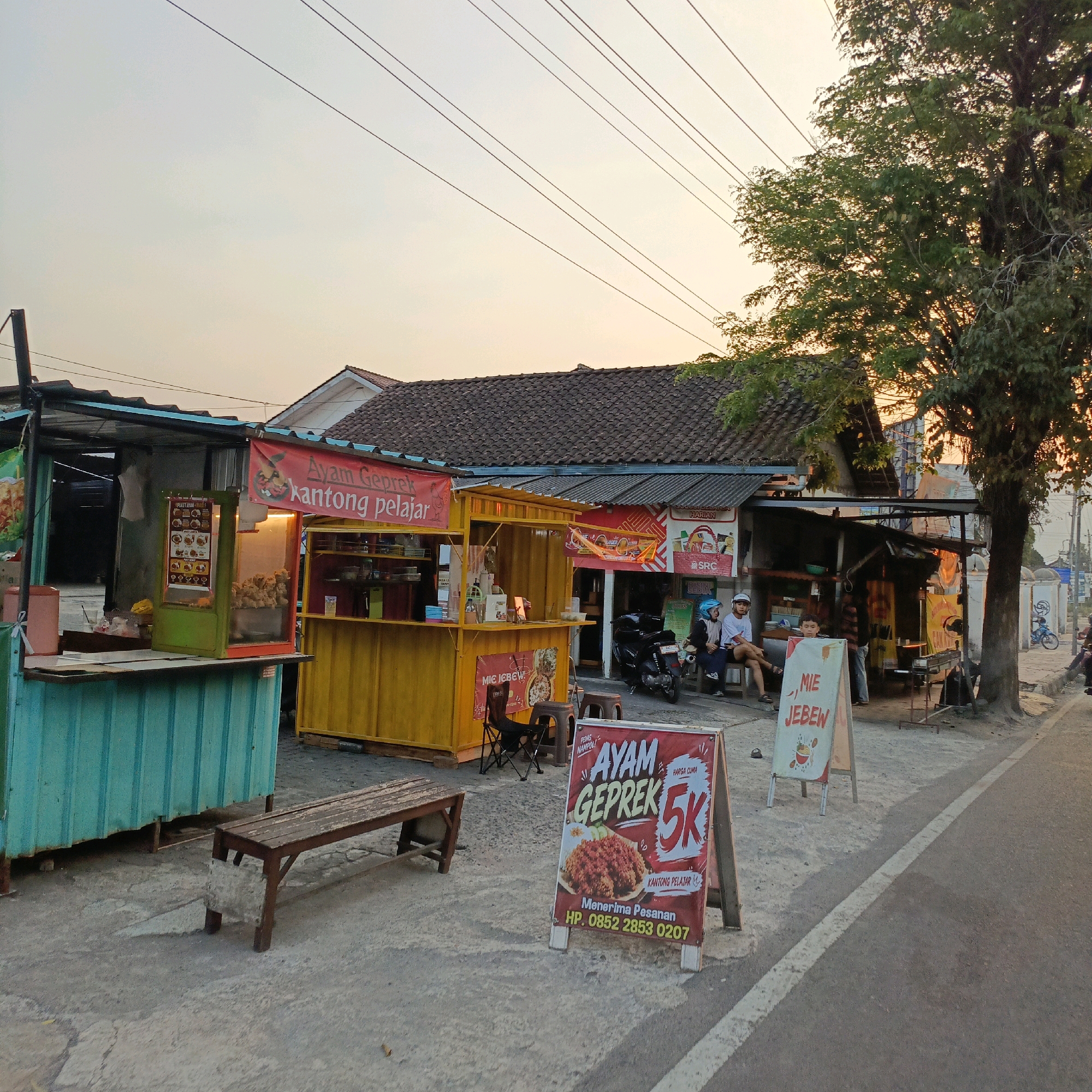 The image depicts a street scene in Salatiga, Indonesia, likely in the late afternoon or early evening due to the soft, warm lighting. Several food stalls are set up along a paved road, with a tree providing shade on the right side.
The closest stall to the left is a teal-colored food cart with a dark roof. It has a counter with items displayed and what appears to be a menu board. Next to it is a bright yellow food stall with a red banner that reads "Ayam Geprek kantong pelajar" (Student's Pocket Fried Chicken). This stall also has a display window showing fried chicken and a menu.
Further down the road, a third stall is visible, with a darker roof and a sign that includes the word "Harian" (Daily). In front of this stall, two people, a man and a woman, are sitting on a bench. The man is wearing a white t-shirt and a blue helmet, and the woman is wearing a white t-shirt and a blue helmet. A motorcycle is parked near them. Another person, a young boy, stands in front of a sign.
A prominent A-frame sign in the foreground advertises "AYAM GEPREK 5K" (Fried Chicken 5000 Rupiah), featuring an image of the dish and contact information. To the right of this sign is another A-frame sign that reads ", MIE JEBEW".
A weathered wooden bench sits in front of the teal food stall. The ground is a mix of concrete and gravel. Power lines crisscross the sky. In the background, traditional Indonesian-style houses with tiled roofs are visible. A blue motorcycle is parked further down the street on the right, near a fence. The overall atmosphere suggests a bustling local food street or market area.