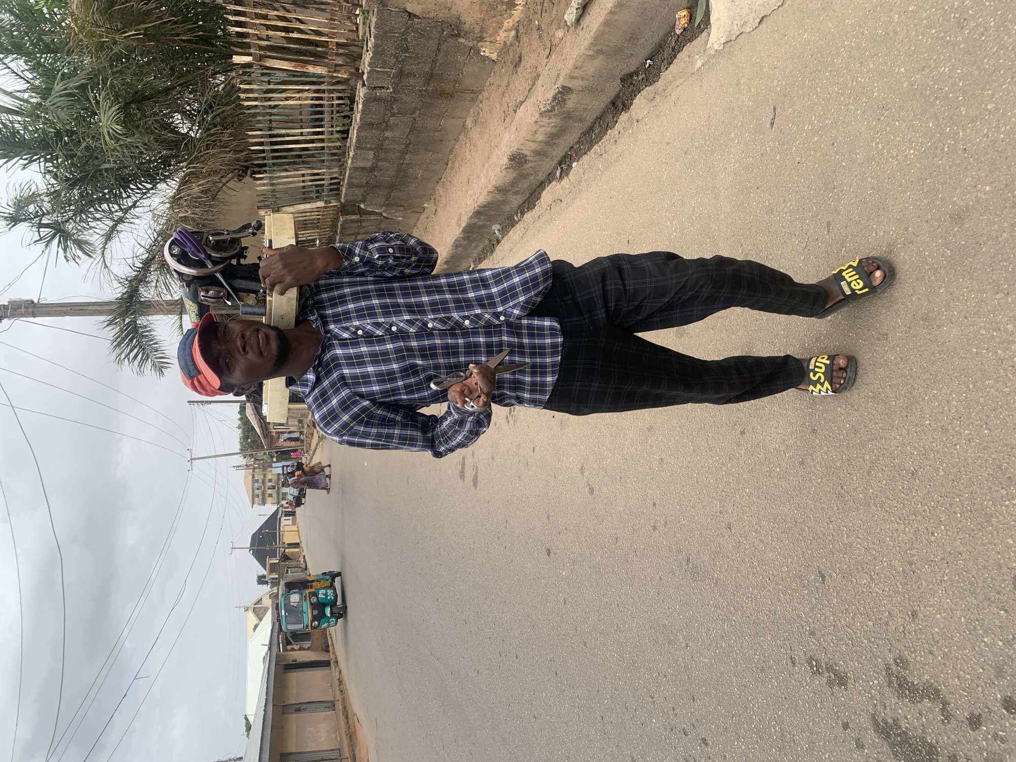 A man stands on an asphalt road in Jos, Nigeria. He wears a red and blue cap, a blue plaid shirt, black pants, and black sandals with yellow designs. He holds a pair of scissors in his left hand and carries a sewing machine on his left shoulder. The street has buildings, power lines, trees, and vehicles like a tricycle in the background. A low concrete wall runs along the side of the road.