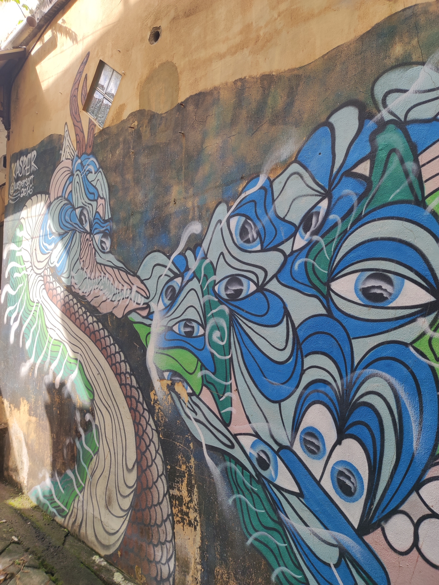 A wall mural in Ubud District, Indonesia, depicts a blue and green serpentine creature confronting a blue eye creature on an old, textured, yellow-brown wall. The wall shows signs of age and wear, with dark discoloration in patches. Above the mural is a small window. The artwork is signed "KASPER @kaspersart 2016". The mural extends to the ground level, which is made of stone tiles.