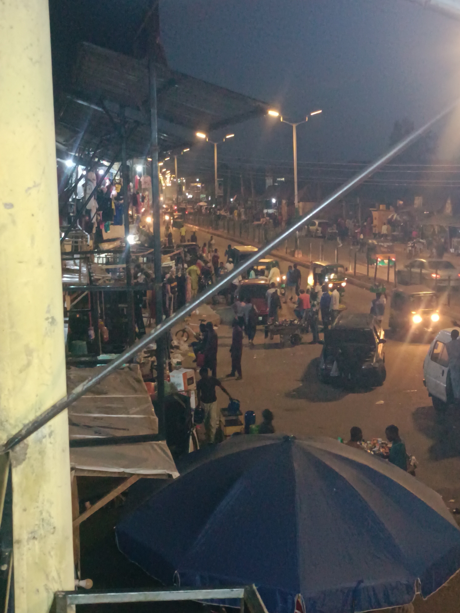 This image captures a bustling night street scene in Bukuru, Nigeria, characterized by vibrant activity and artificial illumination. The photograph is taken from a slightly elevated perspective, looking down onto a street lined with market stalls and numerous people.
The time of day is clearly nighttime, with a dark sky overhead, brightly lit by streetlights and the headlights of moving vehicles. On the far left, a prominent, light-colored concrete pillar occupies the foreground, alongside a dark metal railing that diagonally bisects the image.
The scene is densely populated with people engaged in various activities, suggesting a lively market or commercial area. Individuals are seen walking, standing, and gathering around market stalls. Some figures appear to be vendors attending to their wares, while others are likely shoppers or passersby. On the left side, under a long, dark shelter constructed from metal framing and possibly corrugated sheets, more market activity unfolds, with clothes visibly hanging from racks.
The street is busy with vehicular traffic. Several cars are visible, along with what appears to be an auto-rickshaw or "Keke Napep," and a small white van on the right. Their headlights cast bright glows, illuminating parts of the asphalt and contributing to the dynamic atmosphere.
In the lower foreground, to the right of the central railing, a large, dark blue and white striped umbrella is prominent, indicating another vendor's setup. The overall impression is one of a lively urban environment where commerce and social interaction thrive under the cover of night. No discernible text is visible in the image.