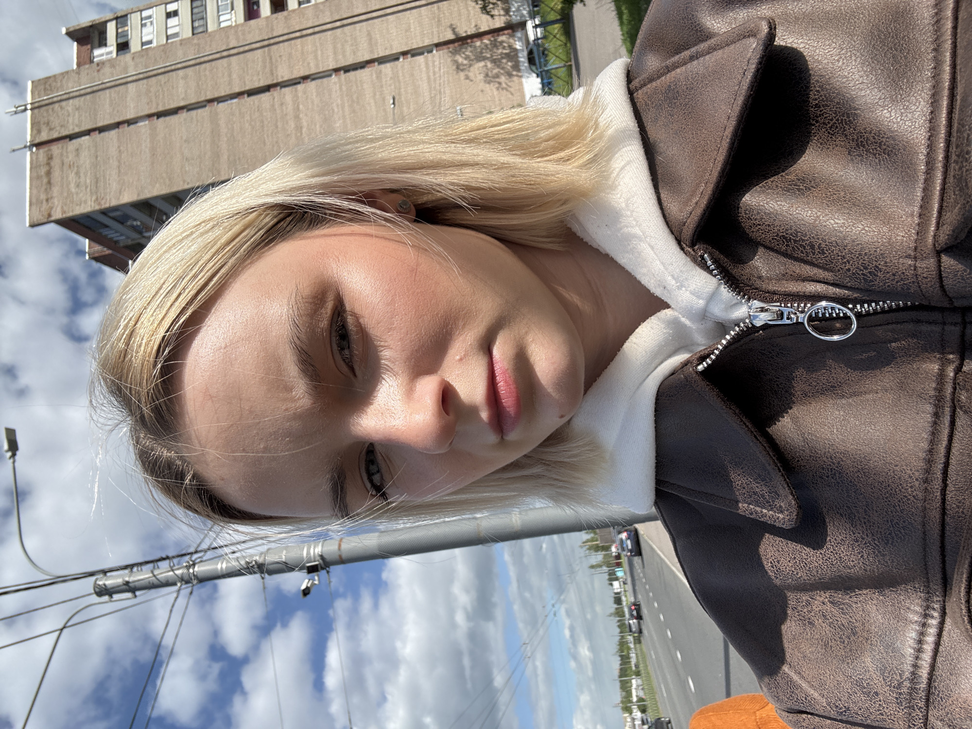 A young woman with light blonde hair, fair skin, and light-colored eyes is pictured in a selfie. She is wearing a brown, possibly faux-leather jacket over a white hooded sweatshirt. The jacket is zipped up, revealing the zipper hardware. The photo appears to be taken outdoors, indicated by the blue sky with scattered clouds and the presence of street features.
Behind her, a multi-story building with a concrete facade is visible. The cityscape includes street lights, a road with vehicle traffic, and glimpses of greenery. The scene seems to be set in Saint Petersburg, Russia.