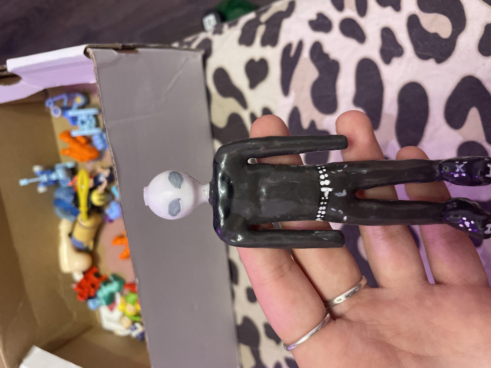 A person's hand with silver rings is holding a black, slender, alien-like figurine with a white head and gray eyes. The figurine is dressed in a black outfit with white dots around the waist and on the boots. In the background, a cardboard box is filled with various small toys, including red, blue, and orange plastic figures, as well as a tan doll. The background surface has a black and white cow-print pattern. The scene is not located in Ladozhskii Trudposelok, Russia.