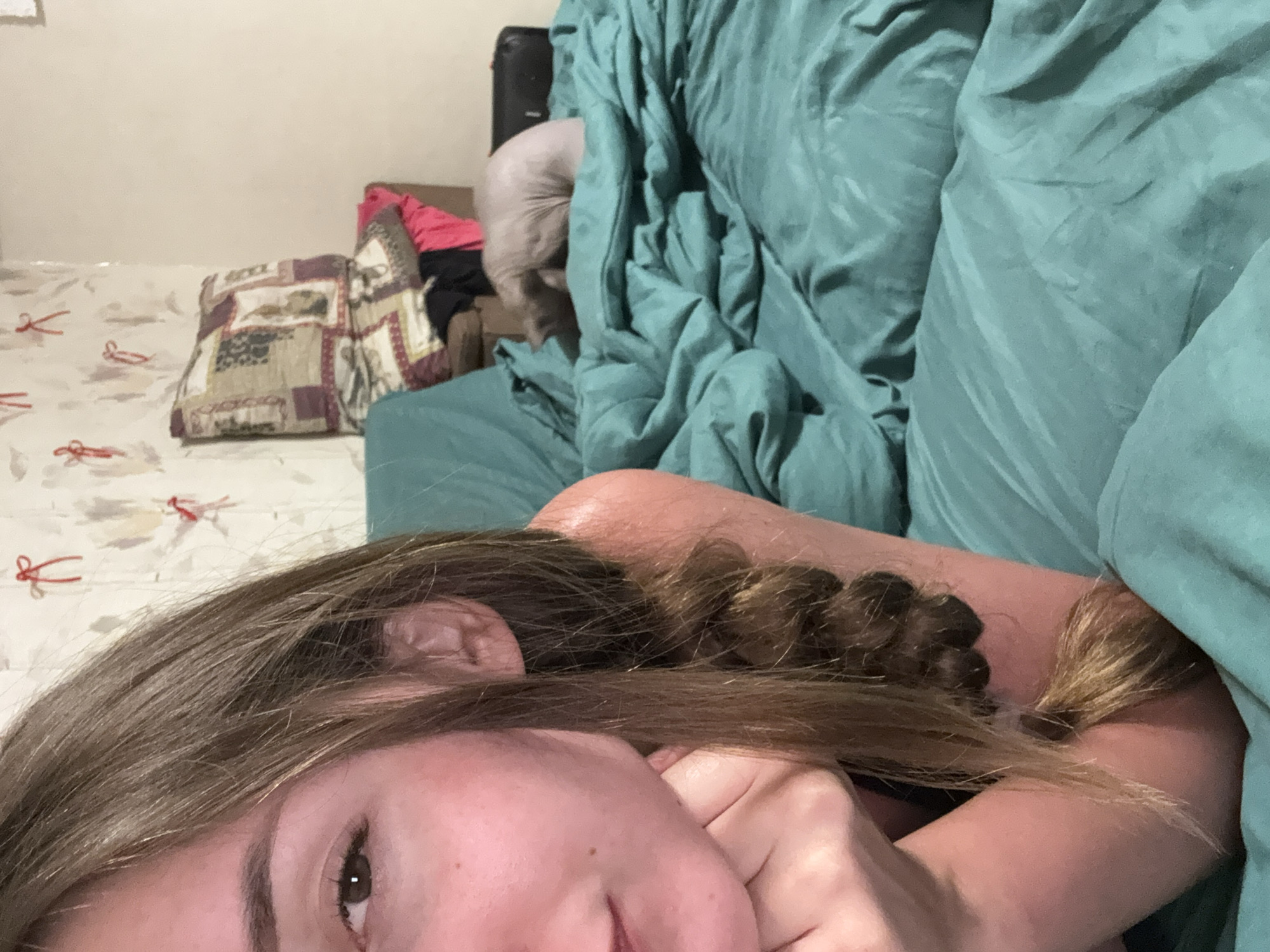 An indoor, close-up, eye-level, slightly angled shot shows a young woman with long brown hair in a braid. Her left arm is bent, with her hand resting on her chin and cheek. She is lying on a teal-colored bed, which is covered with crumpled sheets. In the background, a bald cat is curled up on the bed. The wall behind the bed has light-colored wallpaper with red decorative elements. The room appears to be a bedroom. The image was likely taken in Zhodzina, Belarus.