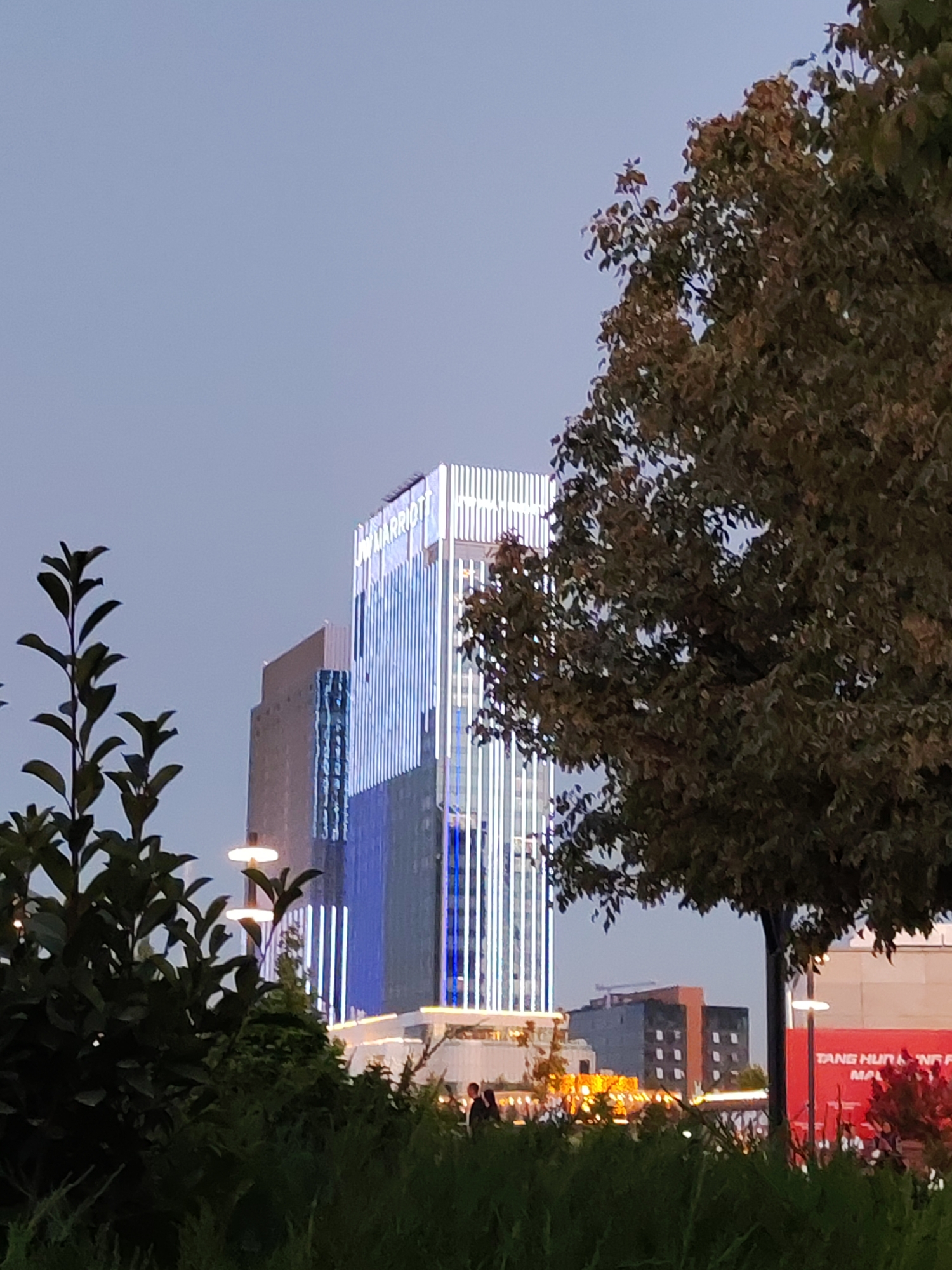 The image depicts a cityscape at dusk, likely in Tashkent, Uzbekistan, with a prominent Marriott hotel building illuminated by blue and white lights. In the foreground, lush green foliage and a large tree with autumnal-colored leaves partially obscure the view of the buildings. The Marriott building, a modern skyscraper, features vertical LED lighting, giving it a striking appearance against the twilight sky. To the left of the Marriott, another building with a grey facade and reflective blue glass is visible. Further in the background, a smaller brick building with a prominent orange facade and white-framed windows can be seen. A large red billboard with partial text, including "TANG HUN NA MA," is positioned on the right side of the frame. Several streetlights with a circular design are scattered throughout the scene. In the lower portion of the image, a few indistinct human figures are present, suggesting public activity in the area. The overall atmosphere is one of urban illumination and natural elements blending together.
